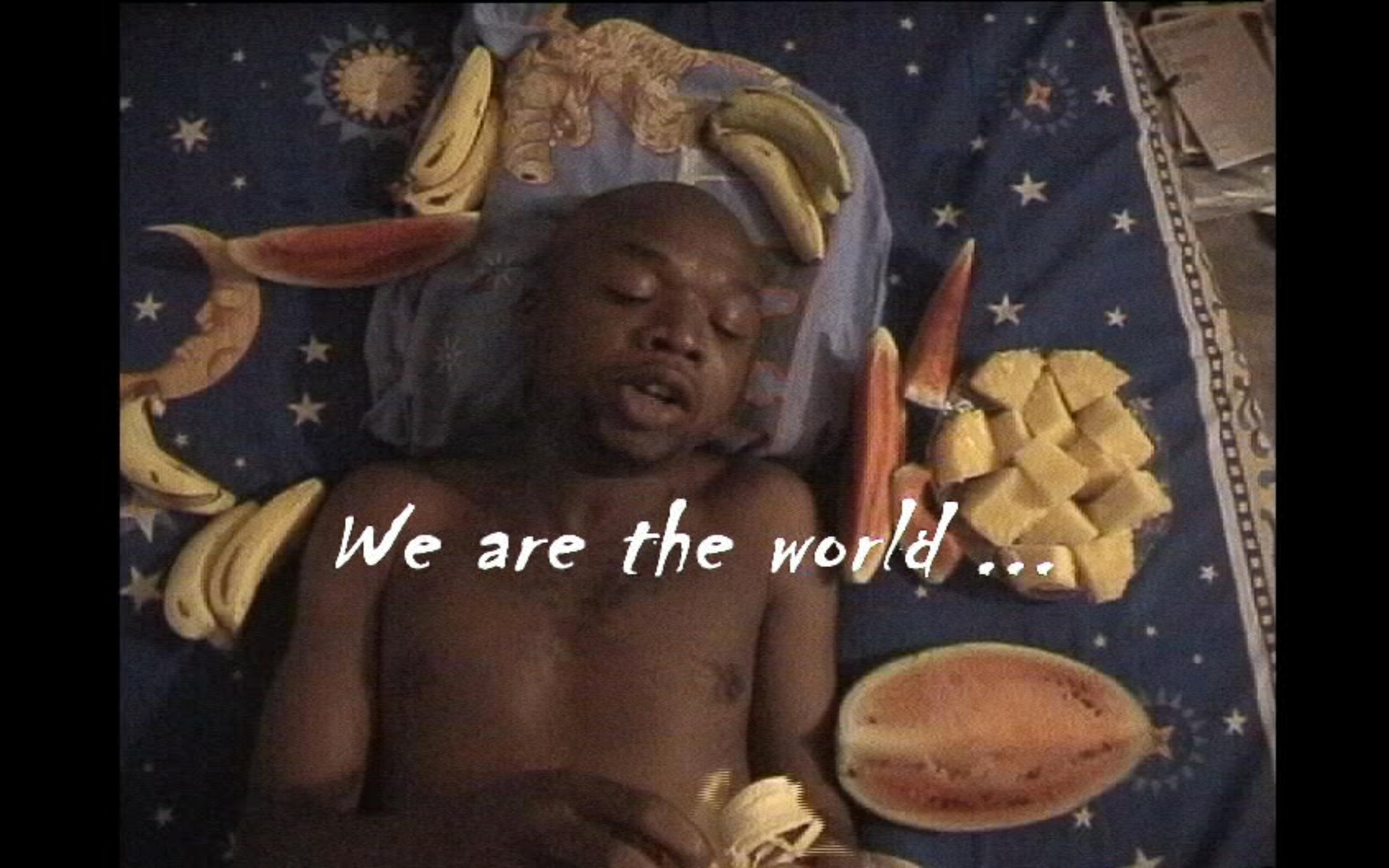 A still frame from Goddy Laye's video 'We are the world' shows an individual lying on fabric, surrounded by pieces of fruit.
