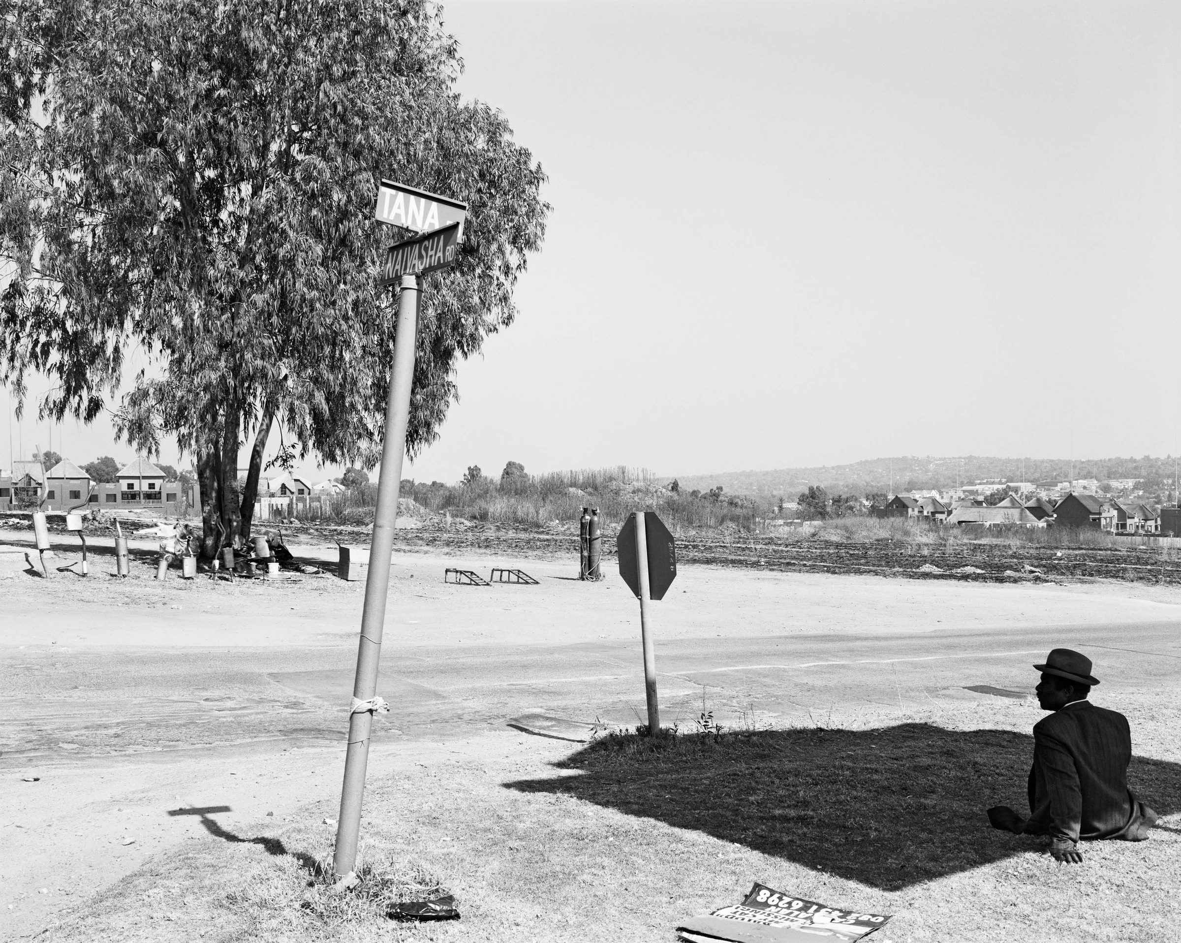 David Goldblatt's black-and-white photograph 'Domestic workers afternoon off, Sunninghill, Sandton' shows a man seated in a spot of shade next to a road. 
