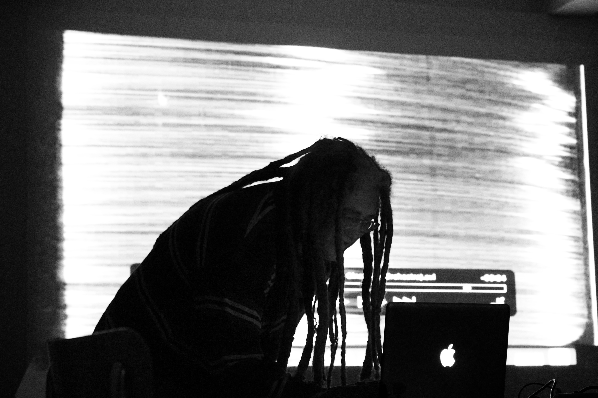 Monochrome event photograph from the ‘Creating in Circles’ exchange on A4’s ground floor. At the front, Eugene Paramoer leans over a laptop. At the back, a video is projected onto the wall.
