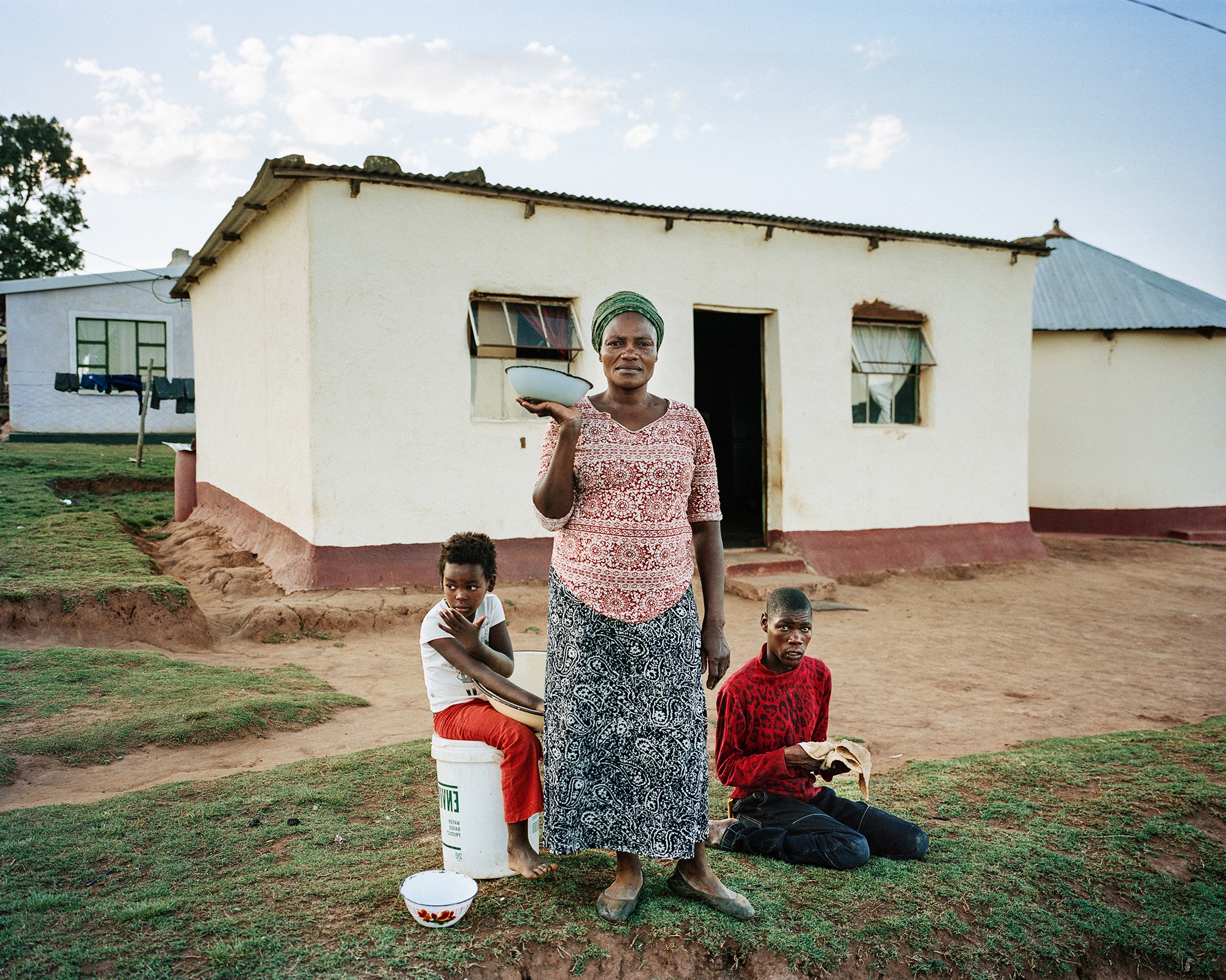 Lindokuhle Sobekwa's photograph 'Ekhwenzane' shows three individuals arranged in front of a square shaped house.

