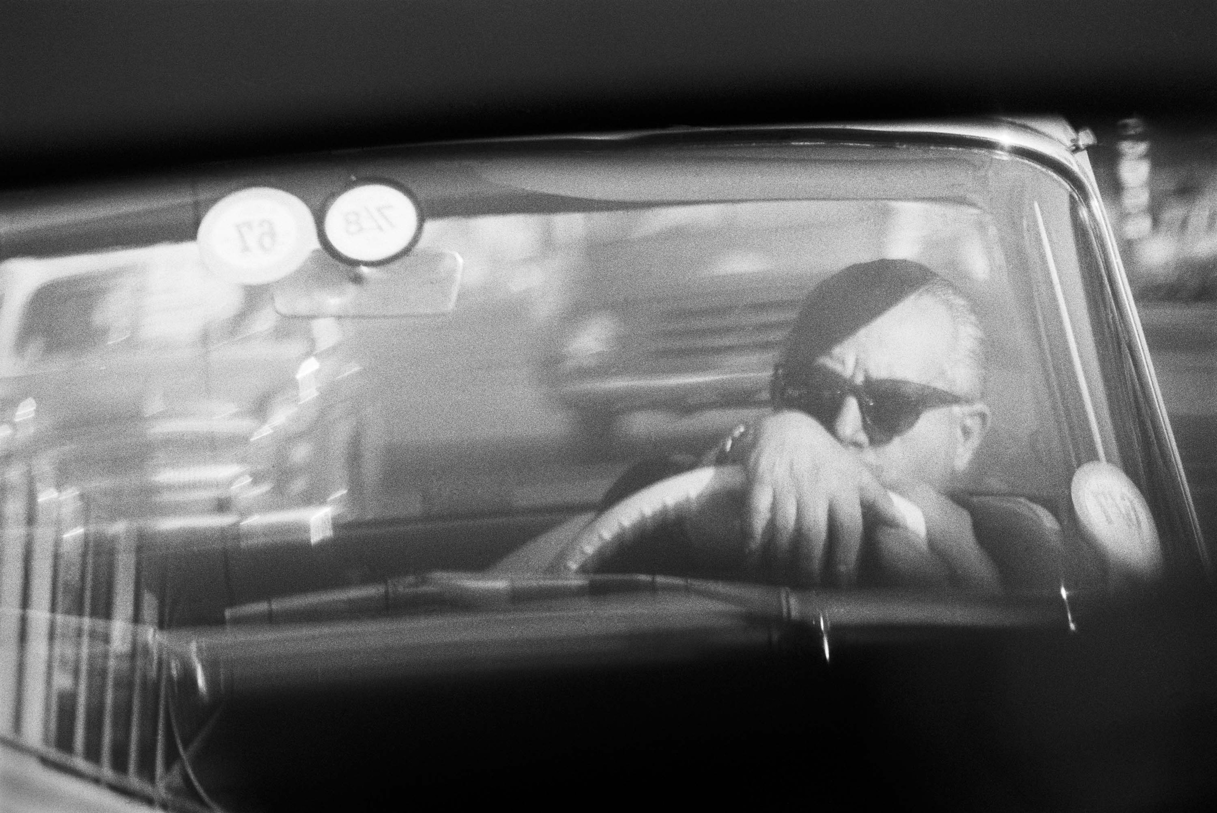 David Goldblatt's monochrome photograph 'Homage to Federico Fellini: while in traffic' shows an individual seated in a car wearing sunglasses.

