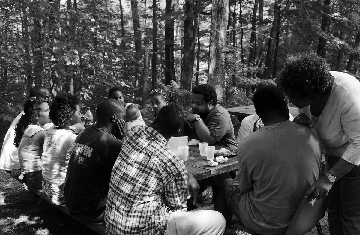 Event photograph from a previous iteration of ‘The Black Lunch Table,’ afterwards hosted on A4’s top floor. Participants are gathered outdoors around a wooden table against the backdrop of a densely wooded area.

