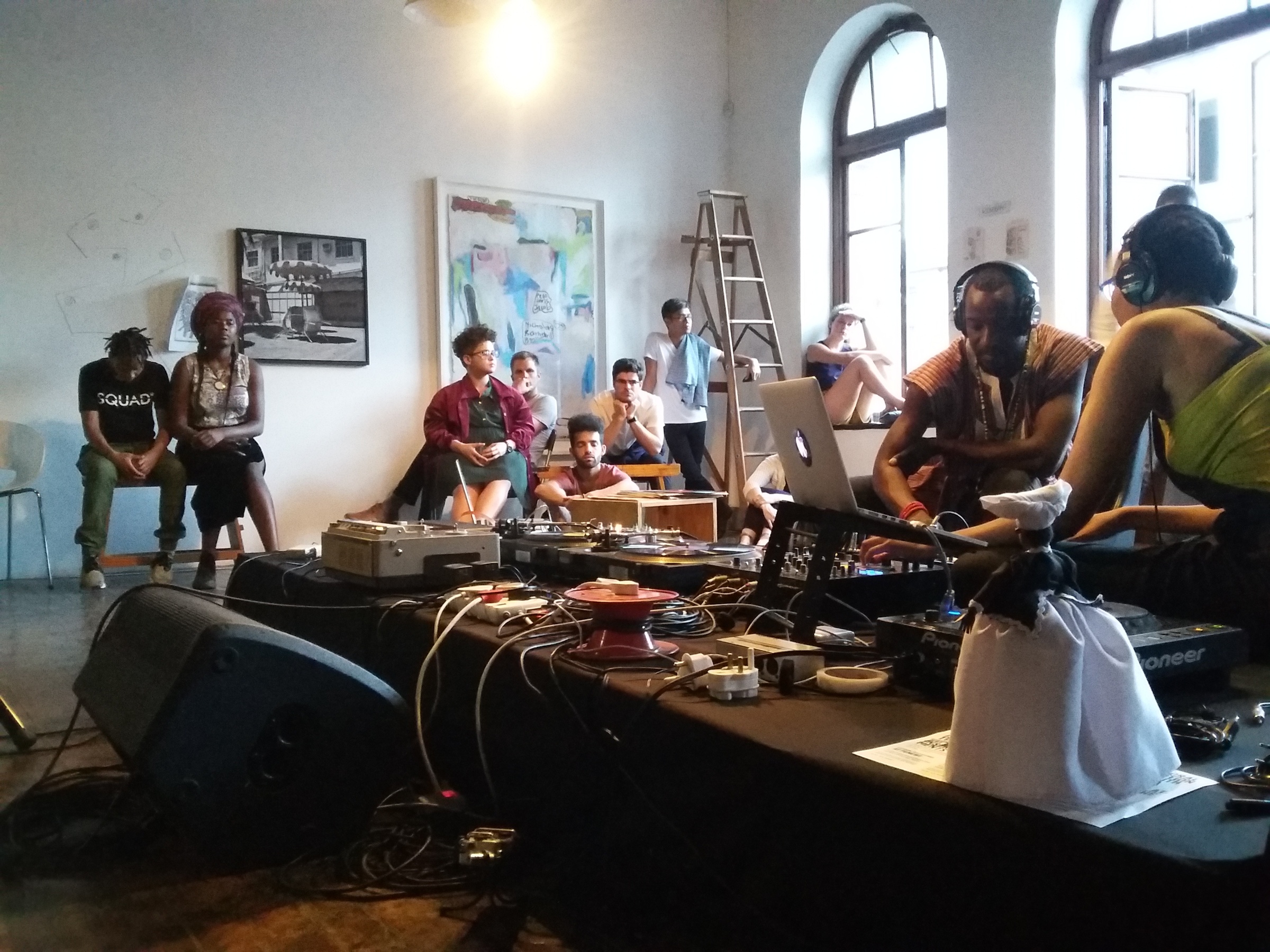 Event photograph from the offsite exchange ‘Pungwe Nights: Listening to Listening’ that shows a table laden with audio equipment is surrounded by various attendees. At the back, Thabiso Sekgala’s photograph ‘The Terrace Hotel, Bulawayo’ and Moshekwa Langa’s mixed media work ‘Untitled (Objects in a Mirror)’ is mounted on the wall.

