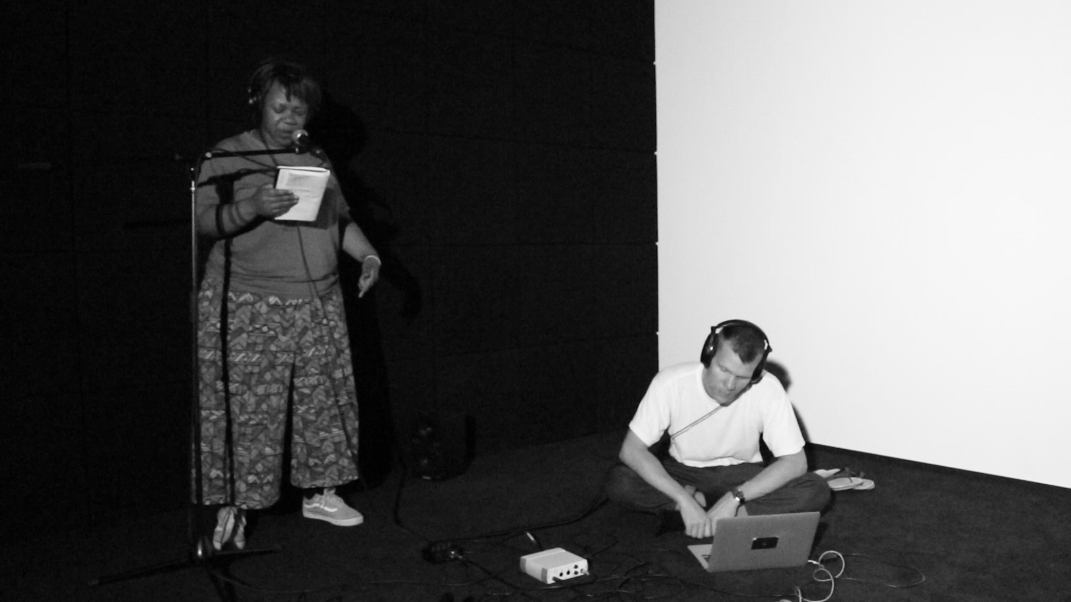 Monochrome process photograph from artists in residence Mushroom Hour Half Hour’s ‘re-vinylize’ sessions on A4’s top floor. On the left, Naledi Chai performs into a standing microphone. On the right, Dion Monti is seated on the floor with headphones and a laptop.
