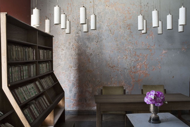 Guy Tillim’s photographic print ‘Library, sports club, Kolwezi, DR Congo’ shows a freestanding bookshelf with printed matter on the left, a table with seating on the right and a time-worn wall at the back.
