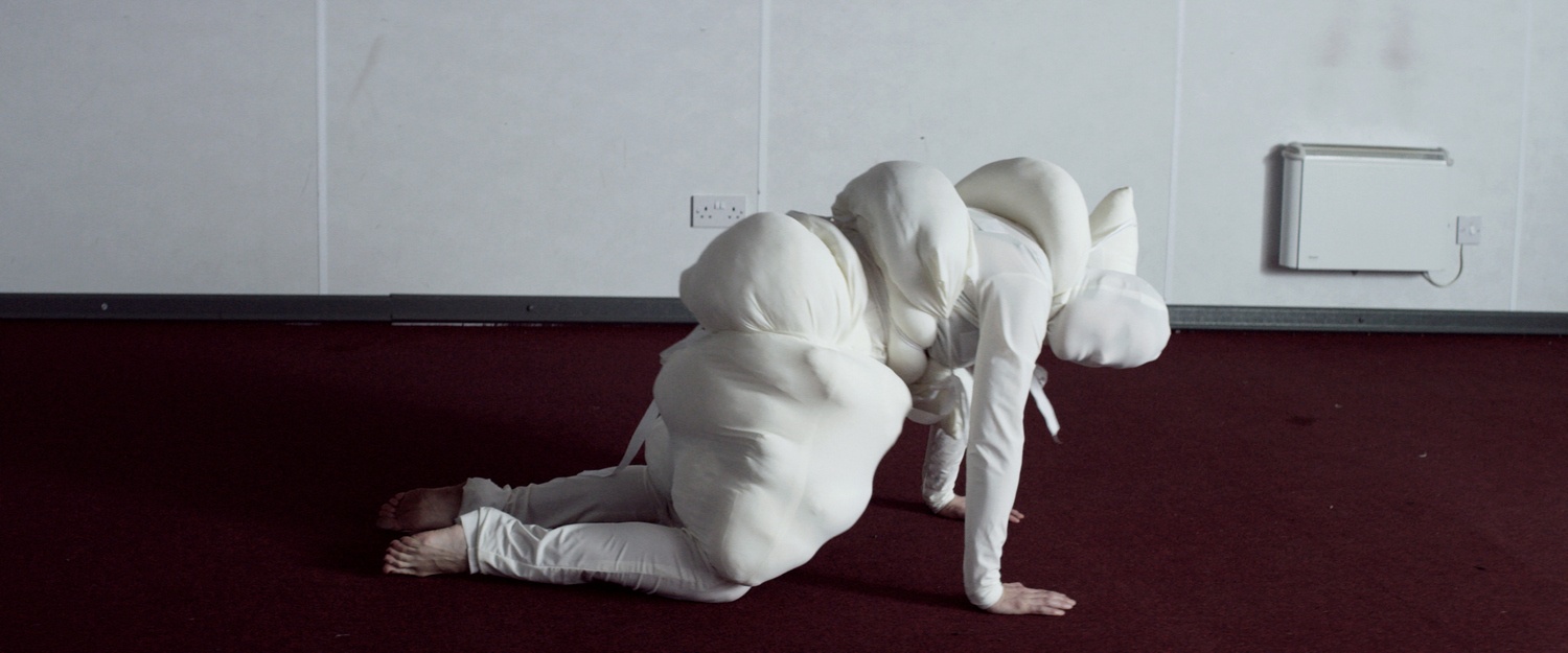 Artwork image from the ‘If I Can't Dance It's Not My Revolution’ exhibition in A4’s Gallery. A still frame from Broomberg & Chanarin’s video work ‘Rudiments’ depicts an individual wrapped in stuffing and white fabric on all fours.
