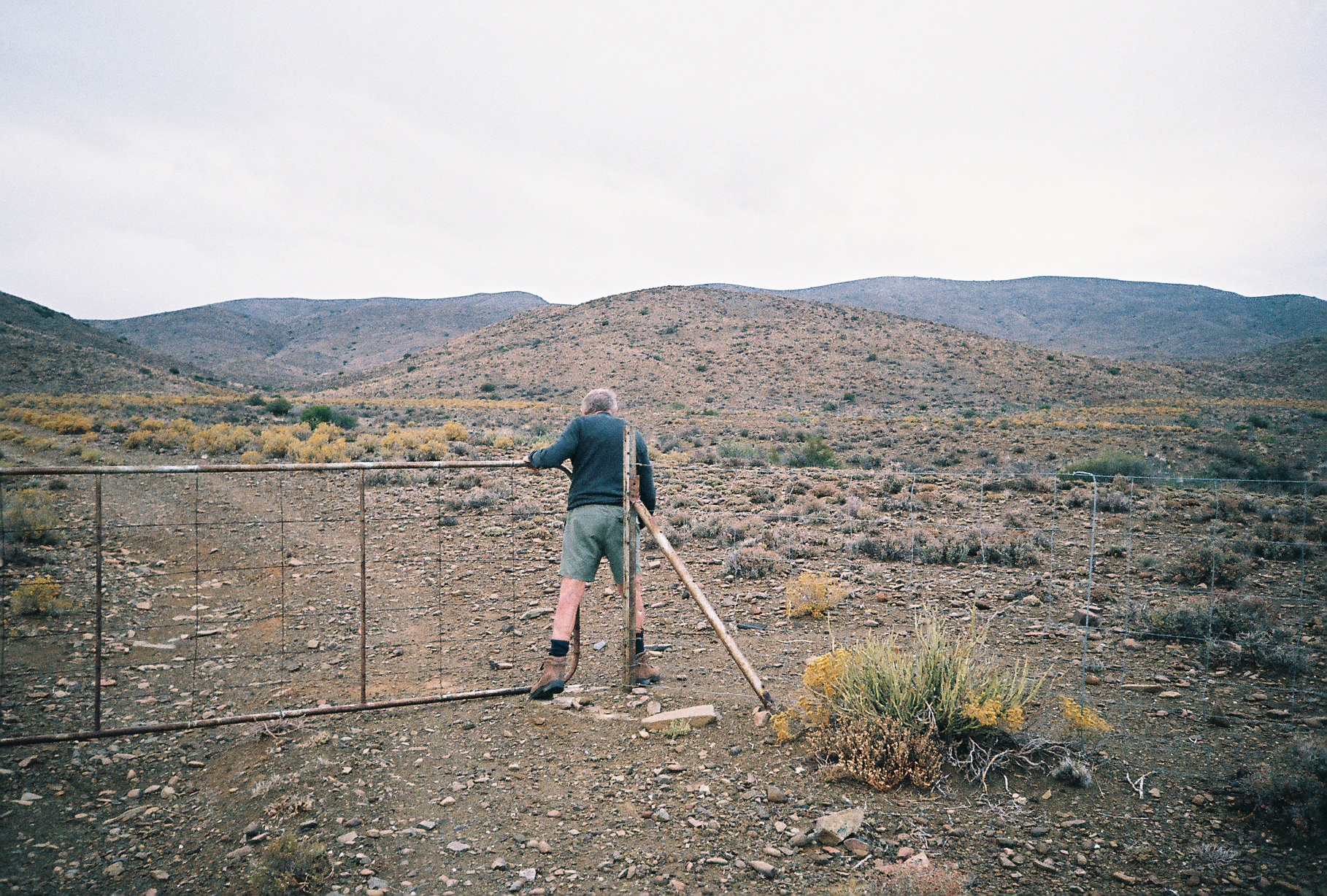 Still frame from the Goldblatt documentary directed by Daniel Zimbler shows David Goldblatt walking through a gate in a wire fence.

