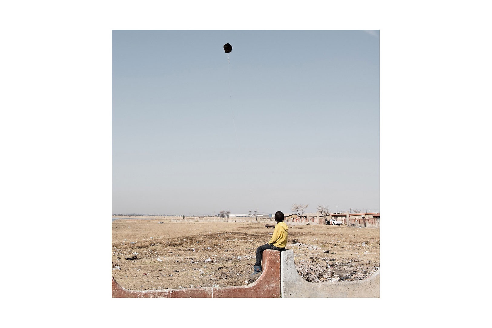 Photograph from Jabulani Dhlamini’s residency on A4’s top floor. At the front, ’Sharpville Kite’ depicts a child sitting on a cement boundary marker flying a kite. At the back, an empty field stretches to the horizon with some houses visible on the right.
