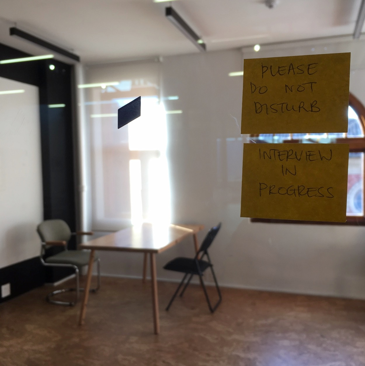 Process photograph from Kathryn Smith’s 2018 artist residency on A4’s top floor. At the front, two notes on A4’s transparent studio door read ‘Please do not disturb’ and ‘Interview in Progress’. At the back, two chairs are arranged around a wooden table.
