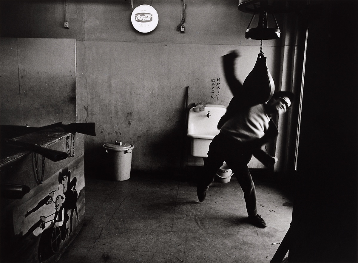 Shōmei Tōmatsu’s monochrome photograph ‘Editor, Takuma Nakahira, Shinjuku, Tokyo’ from the ‘Provoke’ event, a lecture by Matt S Witkovsky on A4’s top floor. On the right, an individual wearing sunglasses punches a pear-shaped punching bag with their back to a washing basin. On the left, a carnival target game is partially visible.
