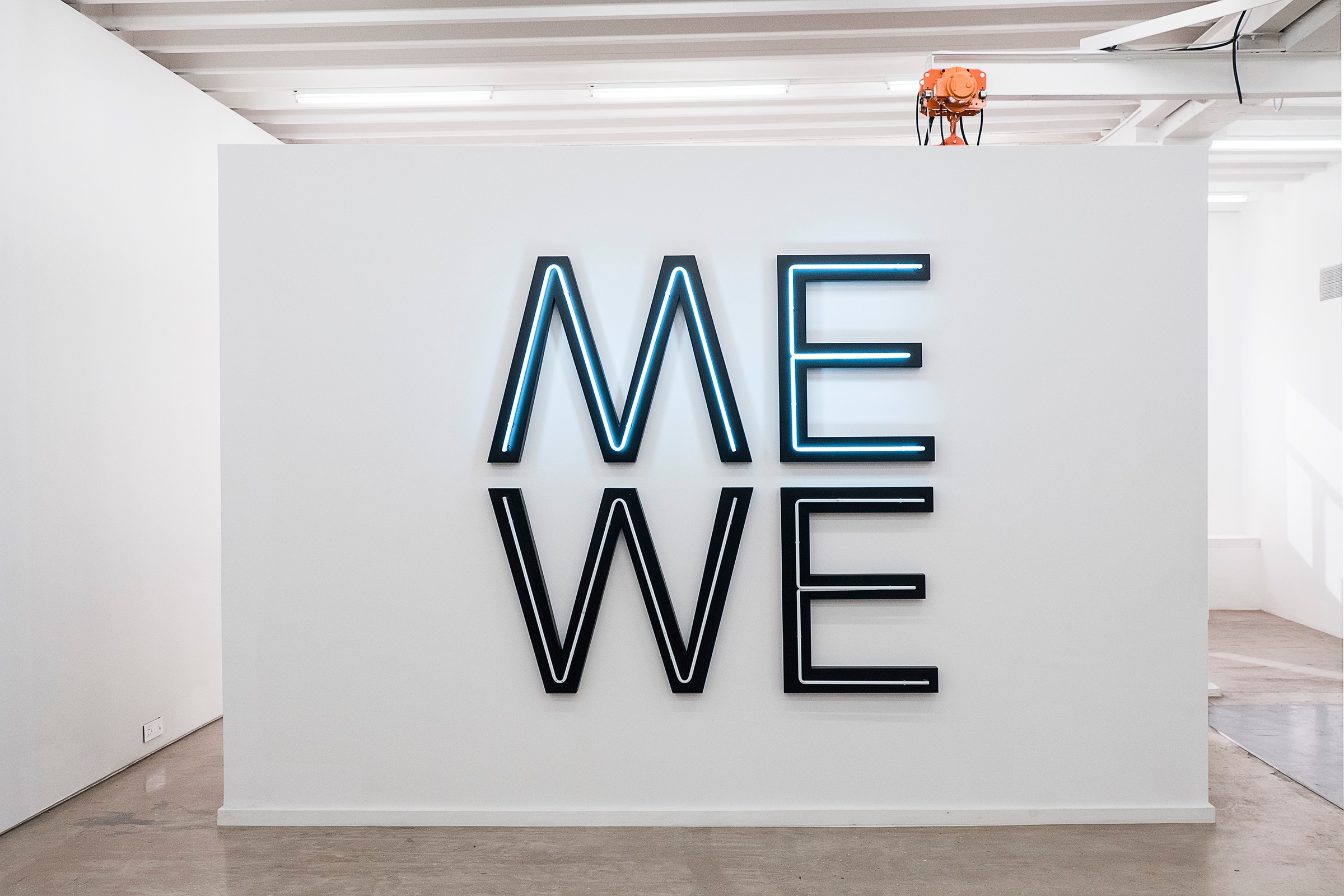 A photograph of Glenn Ligon's black PVC and white neon installation 'Give us a Poem (Palindrome #2)' shows the word 'ME' delineated on a white moveable gallery wall, with the word 'WE' below it.
