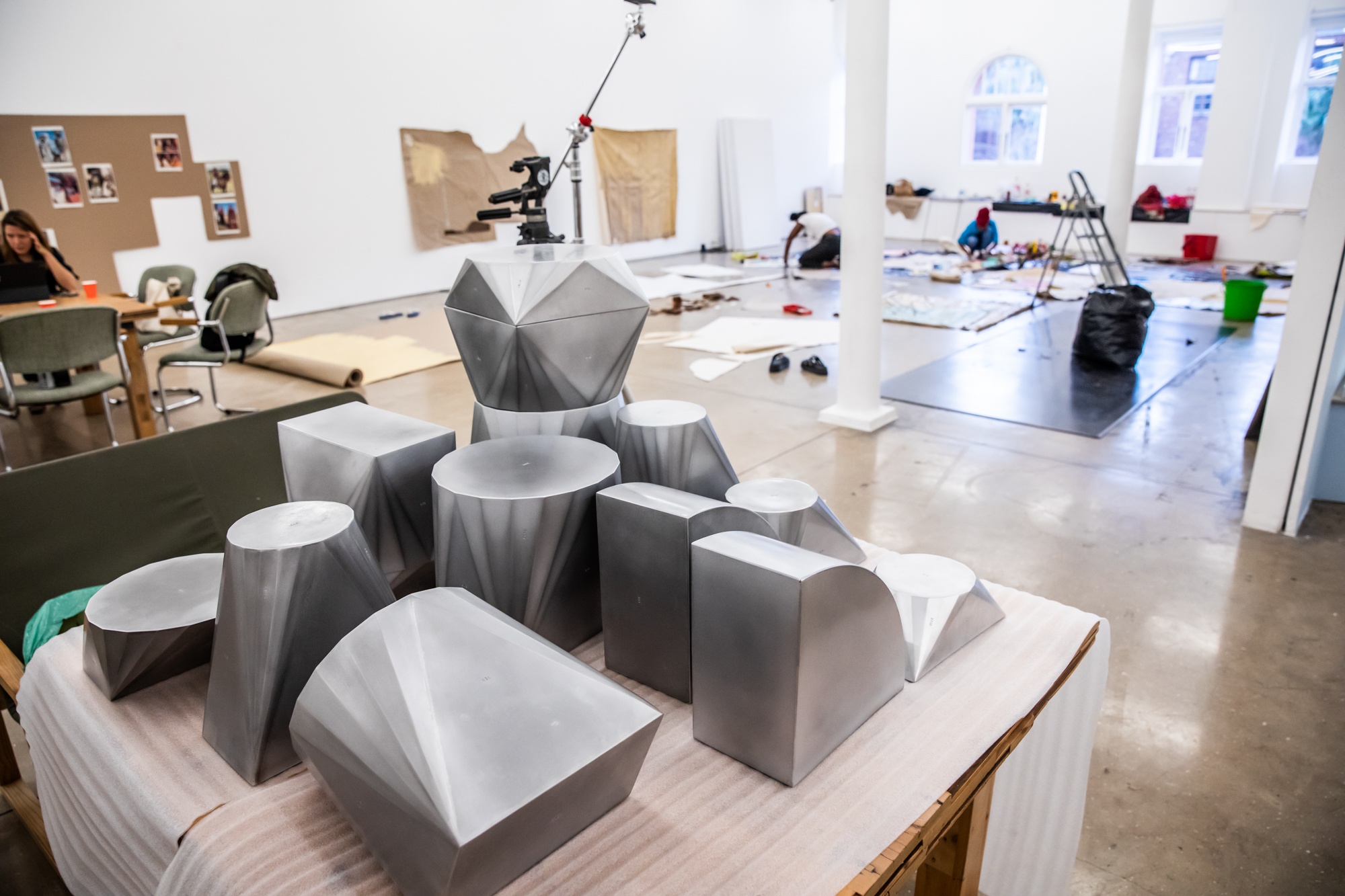 Process photograph from the second rendition of the ‘Parallel Play’ studio setup in A4’s Gallery. At the front, a stack of Kyle Morland’s metal sculptures on a table. At the back, Asemahle Ntlonti and their studio assistant working on the gallery floor.
