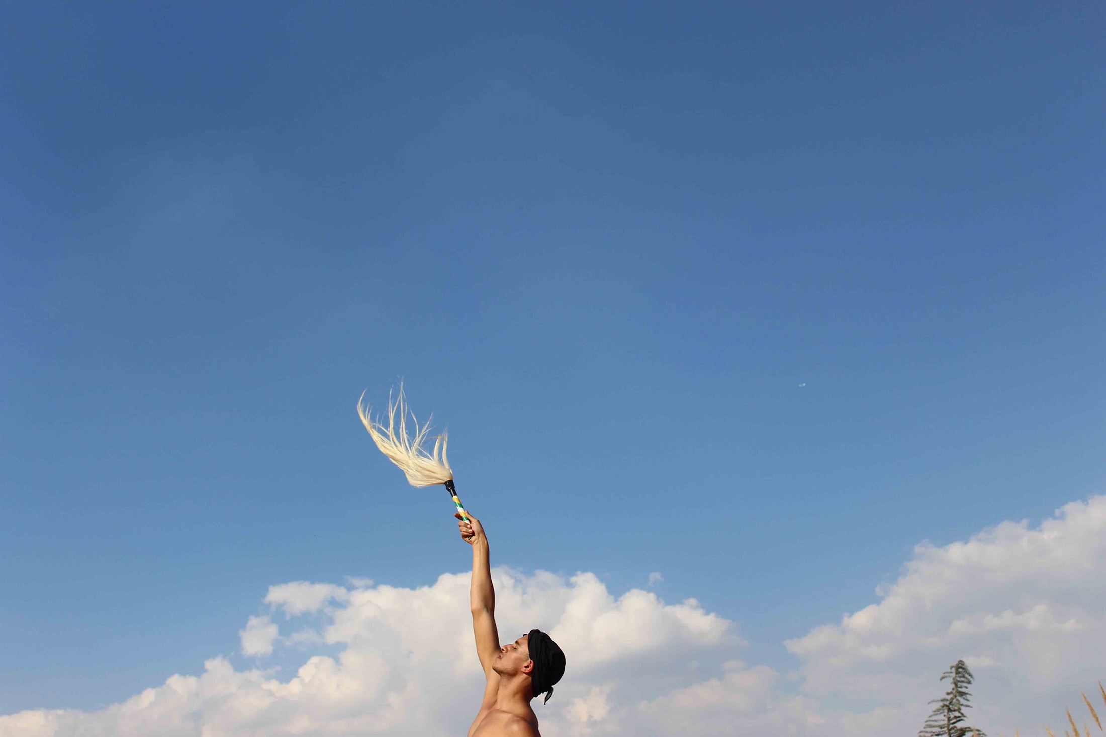 Photographic ephemera from Kieron Jina’s residency at A4. The artist stands against a backdrop of blue sky with one hand raised holding an object resembling a whip.
