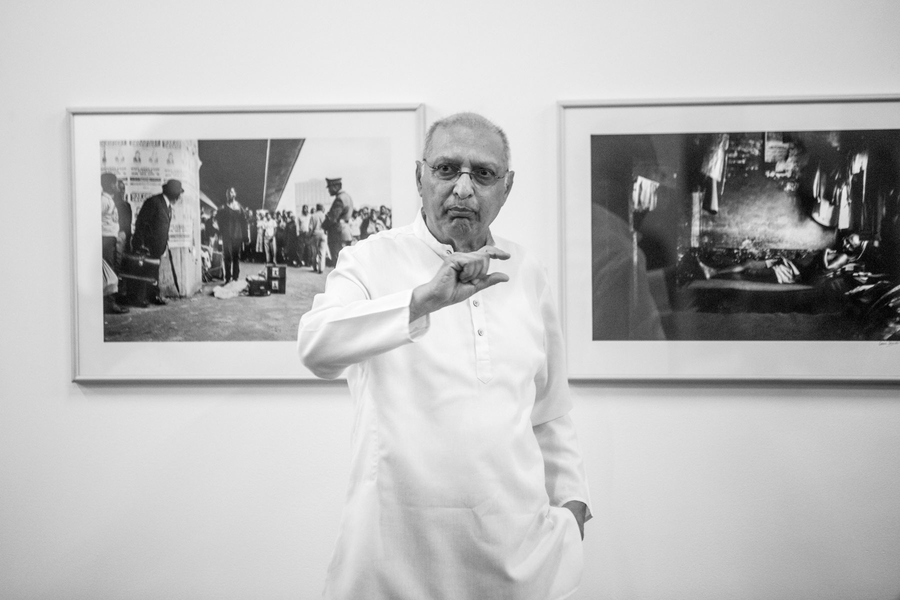 Monochrome event photograph from the launch of Omar Badsha’s book ‘Seedtimes’ on A4’s top floor. At the back, Badsha’s monochrome photographs ‘Street performance, Victoria street, Kwazulu-Natal’ and ‘Migrant worker, Dalton road Hostel, Kwazulu-Natal’ are mounted on the wall. At the front, Badsha stand making a gesture with his right hand.
