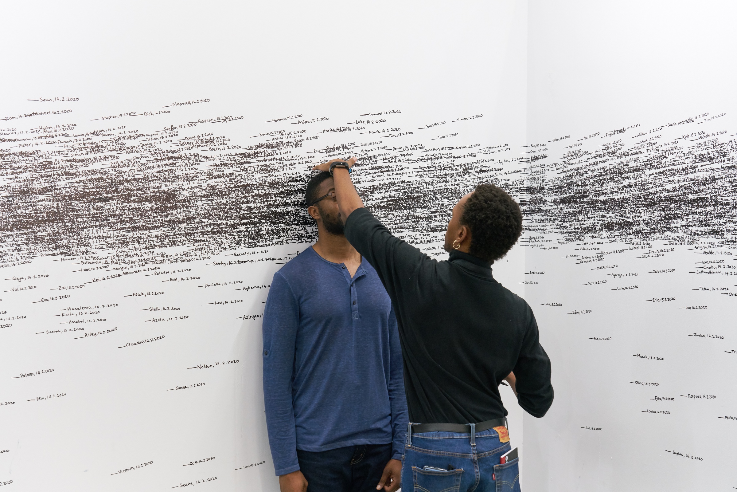 Event photograph of Roman Ondák’s performance piece ‘Measuring the Universe’ at A4’s booth at the 2020 Cape Town Art Fair. At the back, a white wall is covered with overlapping marks that indicate participant’s height, names and the date of their participation in black felt pen. At the front, A4’s Obakeng Motsepe measures the height of a fair attendee.
