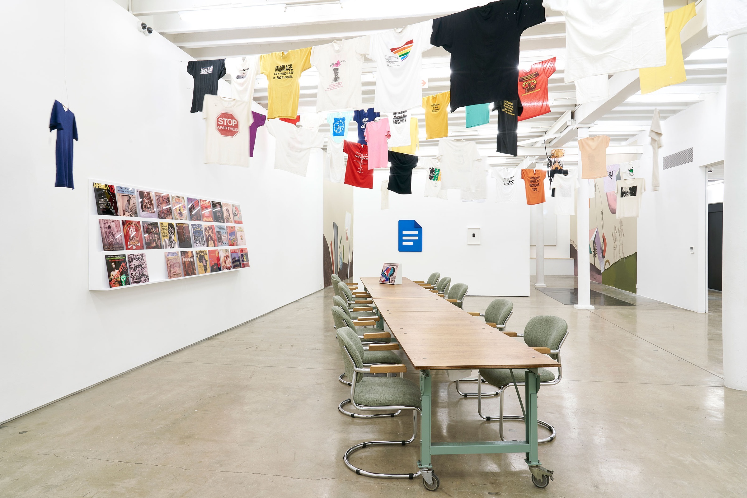 Installation photograph of the Common exhibition. On the left, vinyl record covers from ‘The Library of Things We Forgot to Remember’ by Kudzanai Chiurai are arranged on a slanted white shelf. In the middle is a long wooden table with an interactive box by Mitchell Messina. Above, T-shirts with slogans from the GALA Queer Archive and SAHA are suspended on criss-crossing lines.
