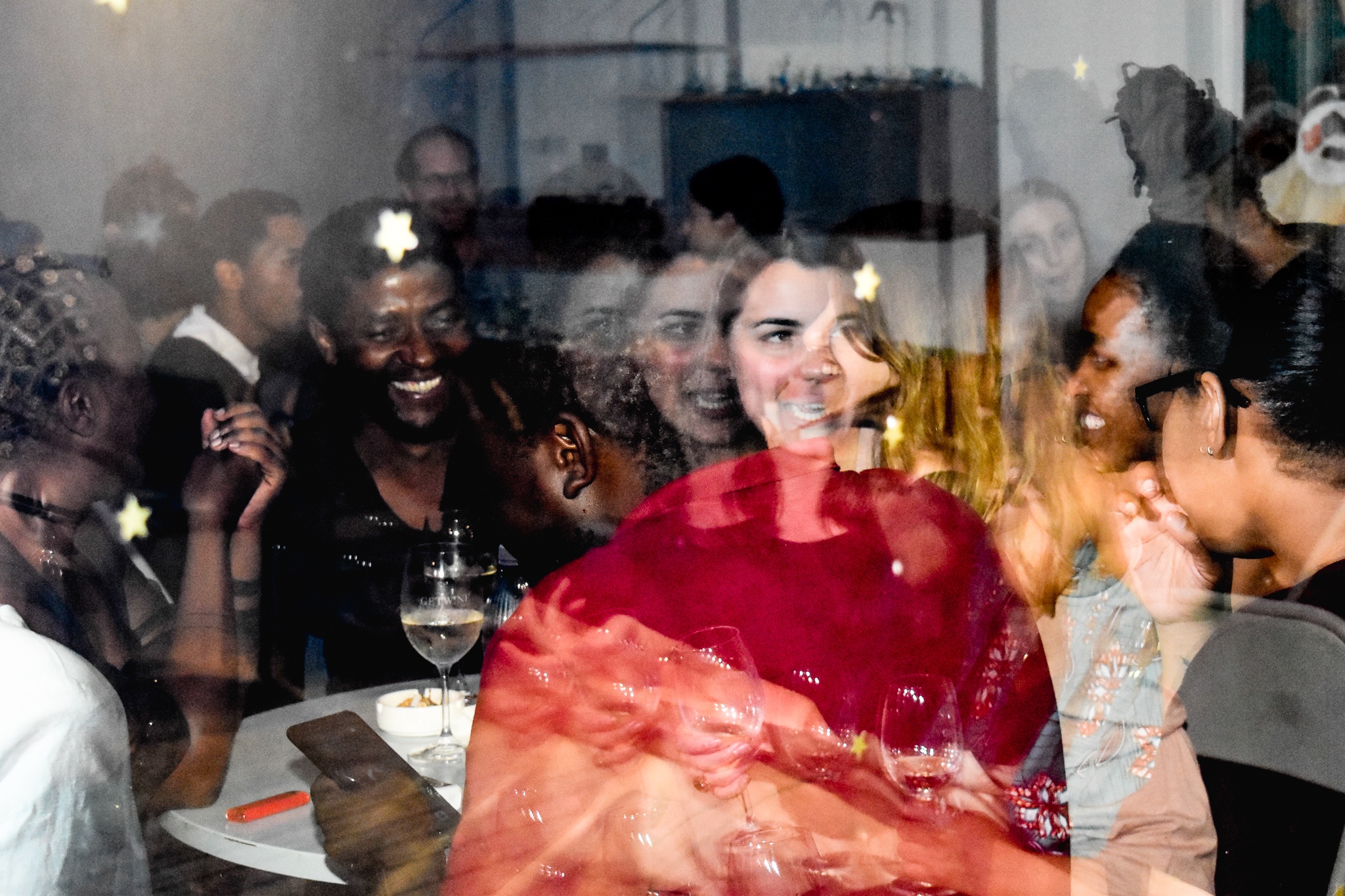 Event photograph from the ‘Sikkum/Sikum’ event on A4’s top floor. At the back, attendees standing in A4’s interior are visible through a glass panel. At the front, attendees sitting at an outdoor table are reflected in the glass panel.
