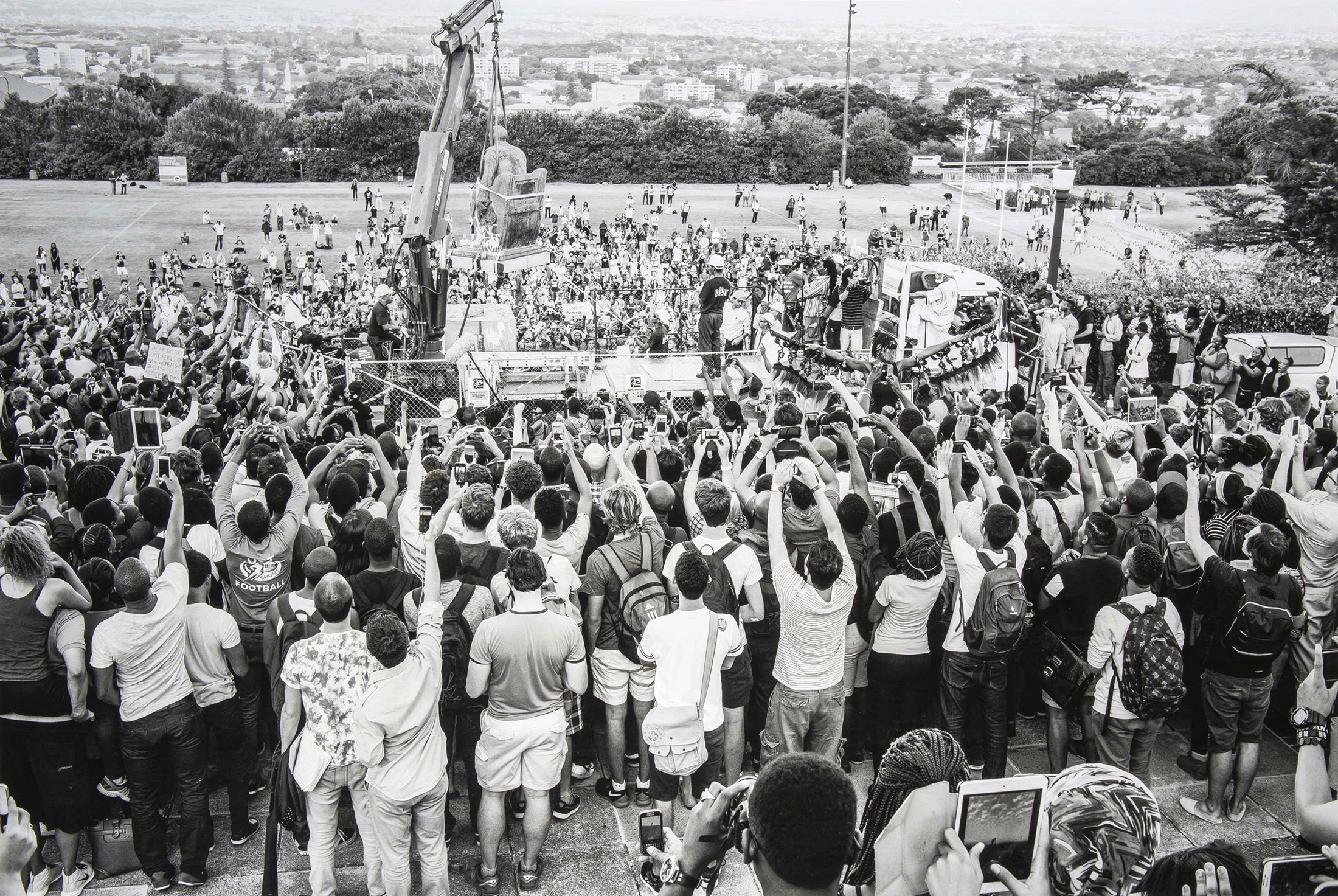 David Goldblatt's black-and-white photograph 'The dethroning of Cecil John Rhodes' shows a statue being lifted off its podium by a crane on a truck. A crowd is gathered in front of the podium with many of the observers holding their cellphones up to photograph the scene.
