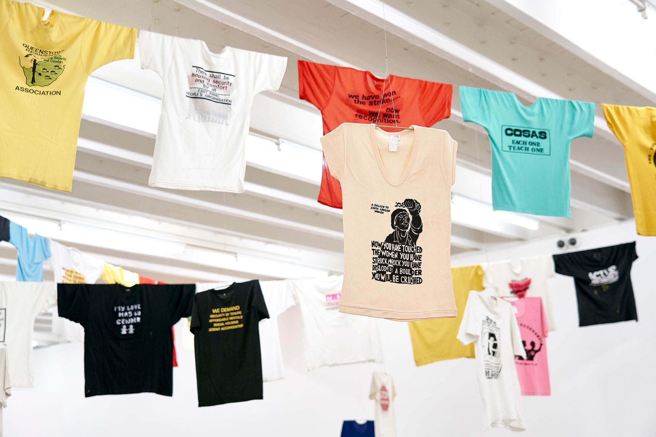 Installation photograph of Common exhibition. Above, t-shirts with slogans from the GALA Queer Archive and SAHA suspended on criss-crossing lines. One beige t-shirt features an image of a shackled woman with raised fist and reads “A Salute to South African Women.”
