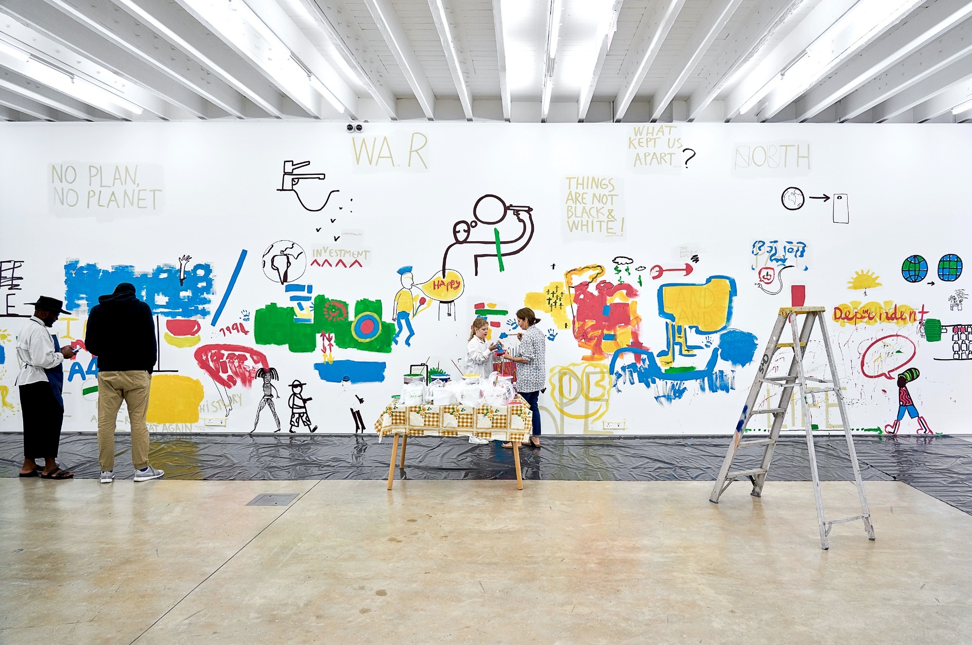 Photograph from the ‘Colourful Erasure’ event in A4’s Gallery. At the front, a table laden with tubs of variously coloured paints and a ladder. In the middle, a sheet of black plastic sheeting lays along the base of the gallery wall with participants standing on top of it. At the back, the gallery wall features monochrome drawings from Dan Perjovschi’s ‘Black & White Cape Town Report’ exhibition overlain with colourful drawings by participants.
