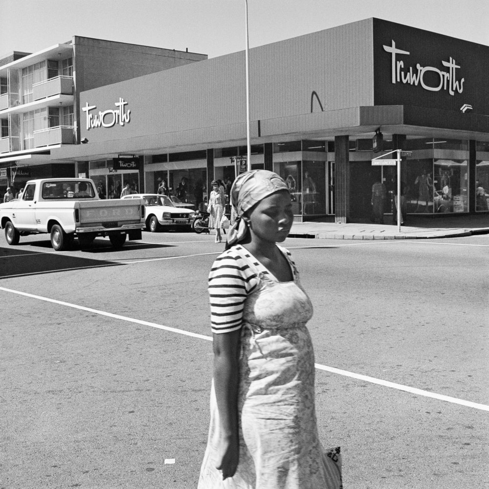 David Goldblatt's monochrome photograph 'On the corner of Commissioner and Eloff Streets' shows a standing individual at the front, with a clothing shop across the street at the back.
