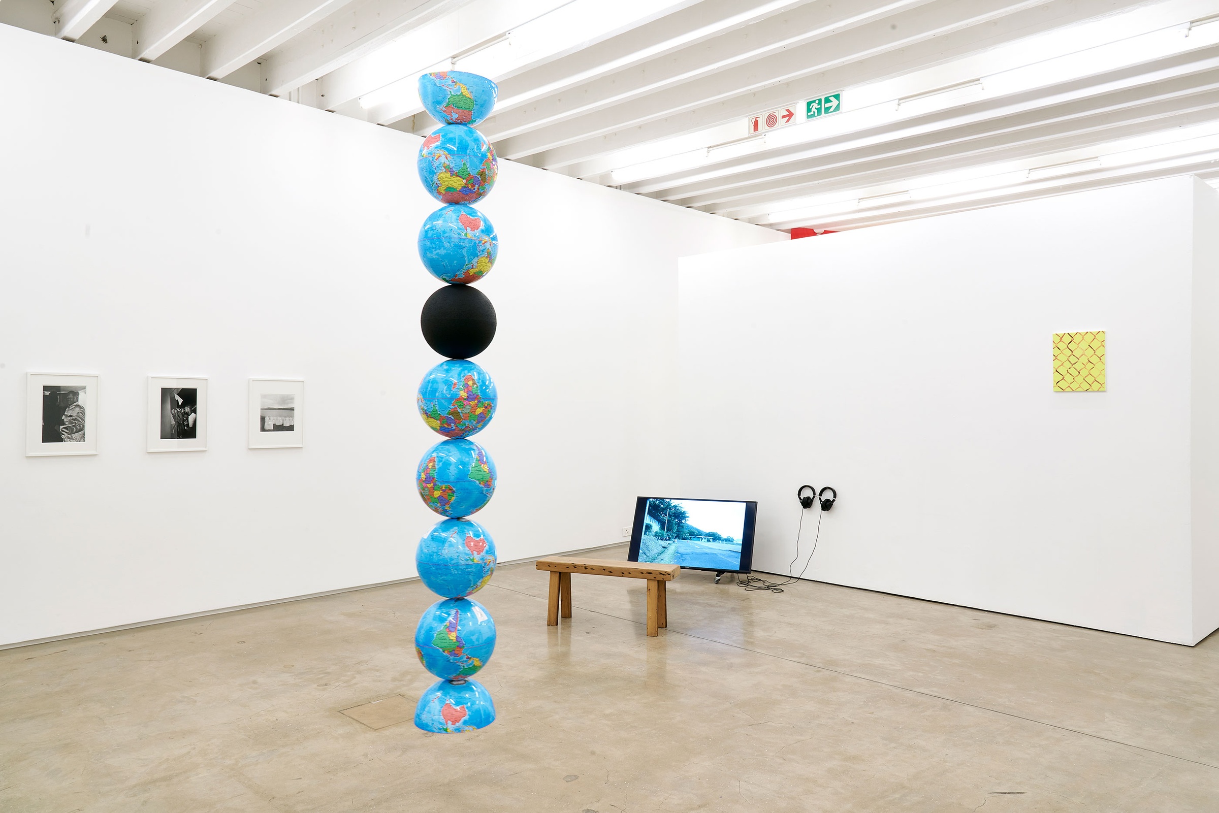 Installation photograph of Common exhibition. To the left, Nolan Oswald Dennis’ ‘model for an endless column,’ a column of globe models suspended from the ceiling. One is solid black, the first and last are halved. On the right, a video screen playing Francis Alÿs’ ‘Painting/Retoque’ sits on the floor, with earphones and bench.
