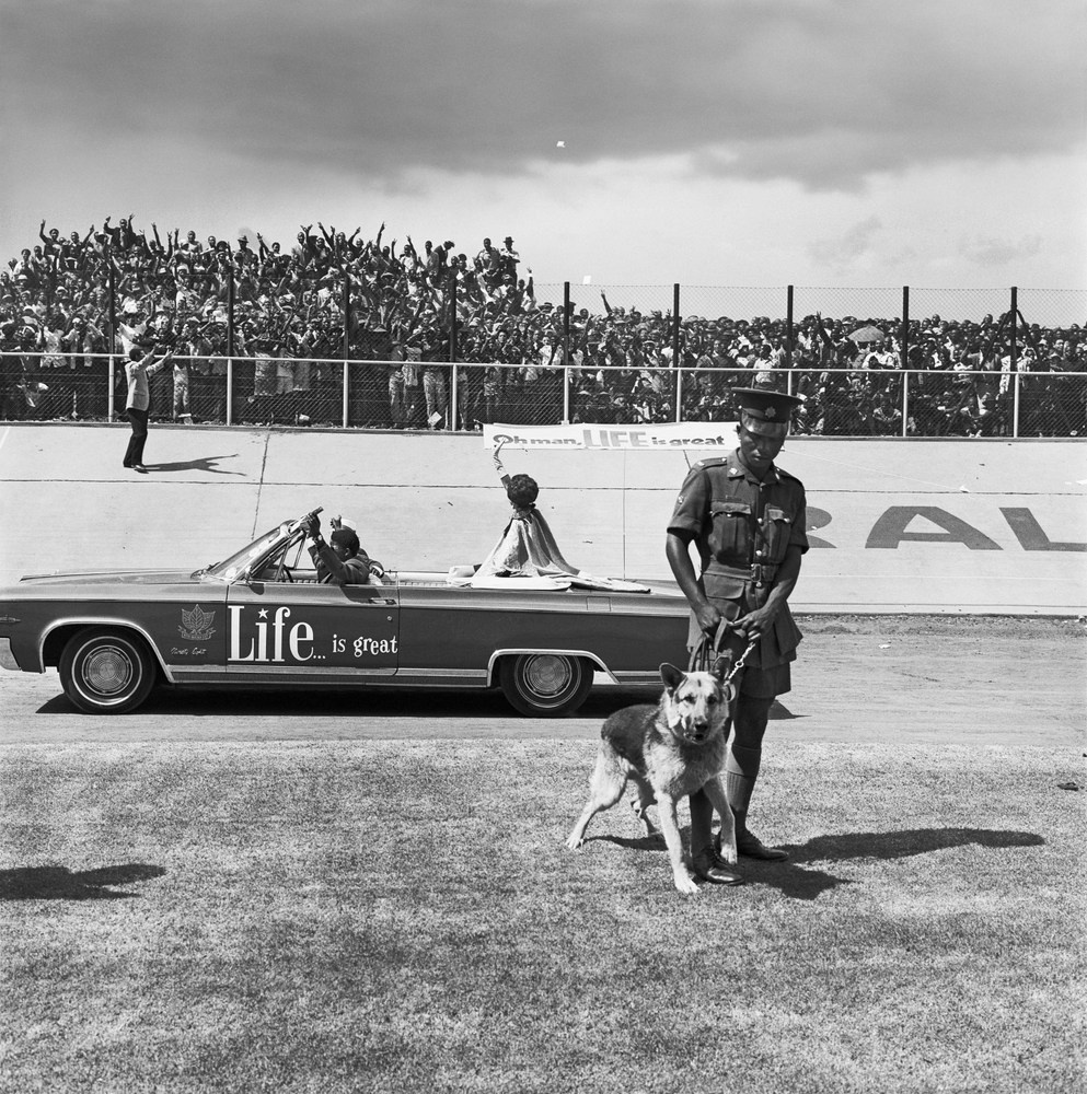 David Goldblatt's monochrome photograph  'Cup Final, Orlando Stadium, Soweto. 1972', depicts a uniformed figure restraining a dog in front of a parade in a stadium.
