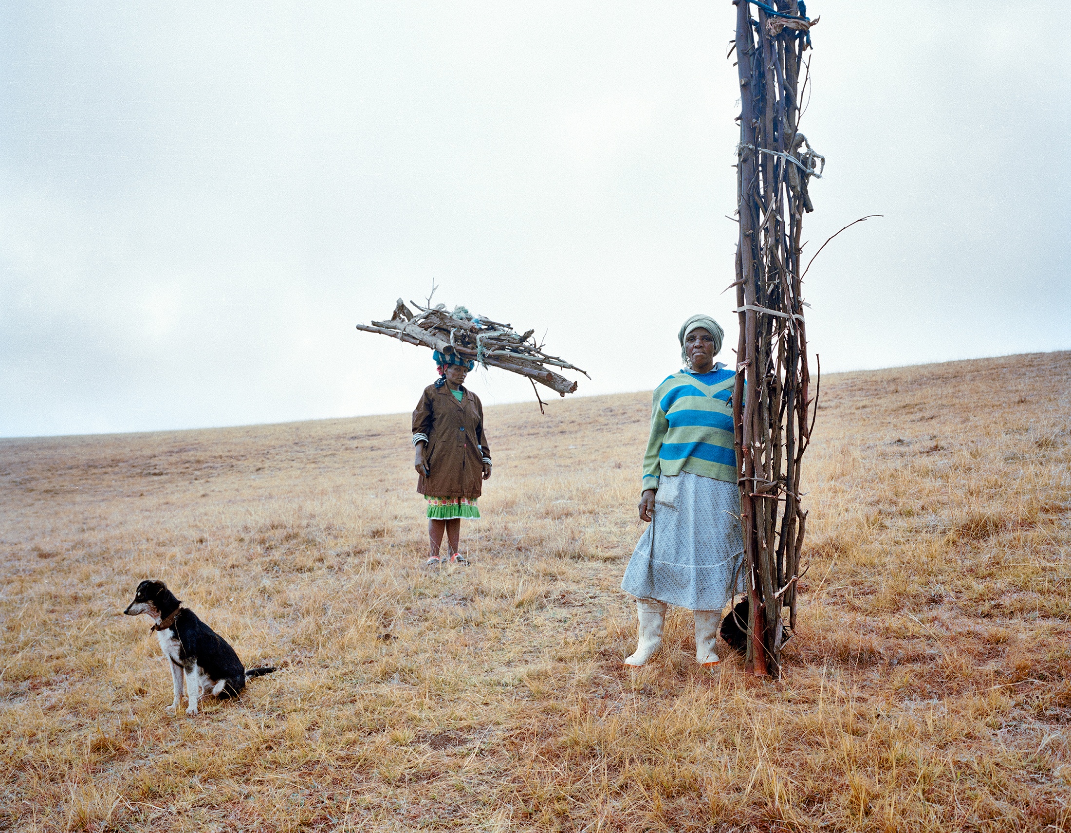 Lindokuhle Sobekwa's photograph 'Omama bathwelinyanda' shows two individuals holding long bundles of sticks in a grassy plain.
