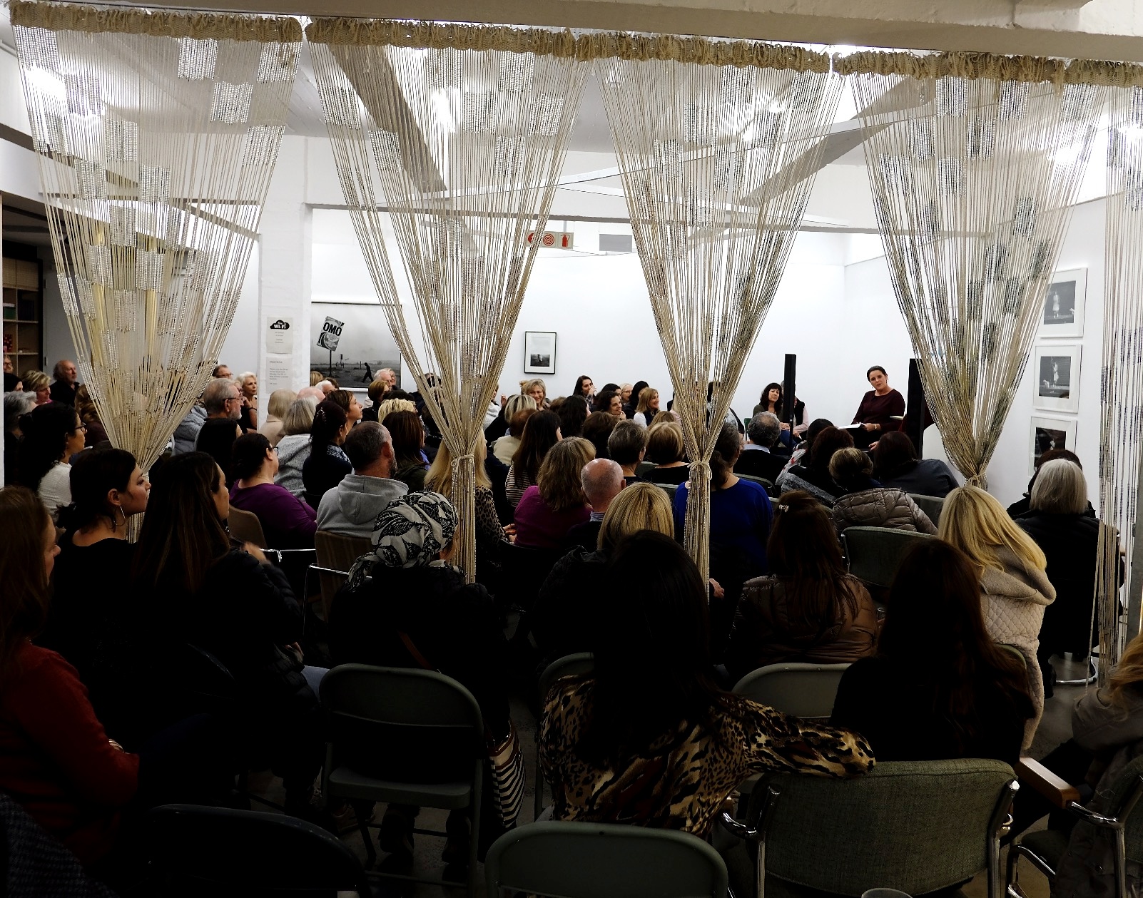 Event photograph from a conversation with Heather Morris, author of ‘The Tattooist of Auschwitz’, on A4’s ground floor. The room is filled with seated attendees, with the speakers seated along the right-hand wall. Igshaan Adams’ mixed media tapestry ‘Al asma ul Husna’ is suspended from the ceiling, bunched to allow space for the crowd.
