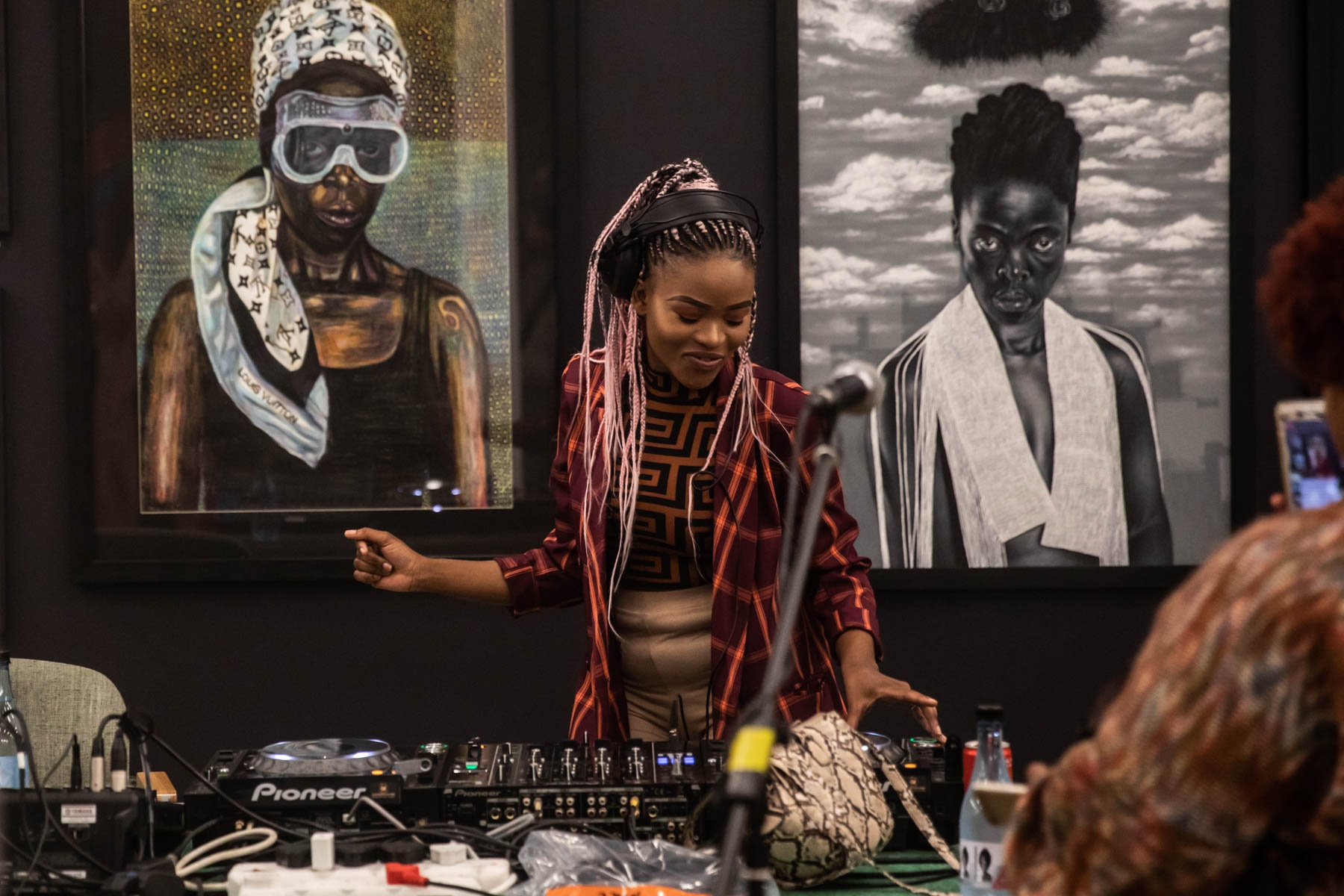 Event photograph from ‘Finissage: Ikhono LaseNatali’ in A4’s Gallery. At the front, DJ Portia Luma operates a turntable. At the back, two artworks are hung on a black wall. On the lefthand side, Mthobisi Maphumulo’s oil pastel drawing ‘Somnyama Ngonjama.’ On the righthand side, Mduduzi Dzanibe’s acrylic painting ‘Somnyama Ngonyama.’
