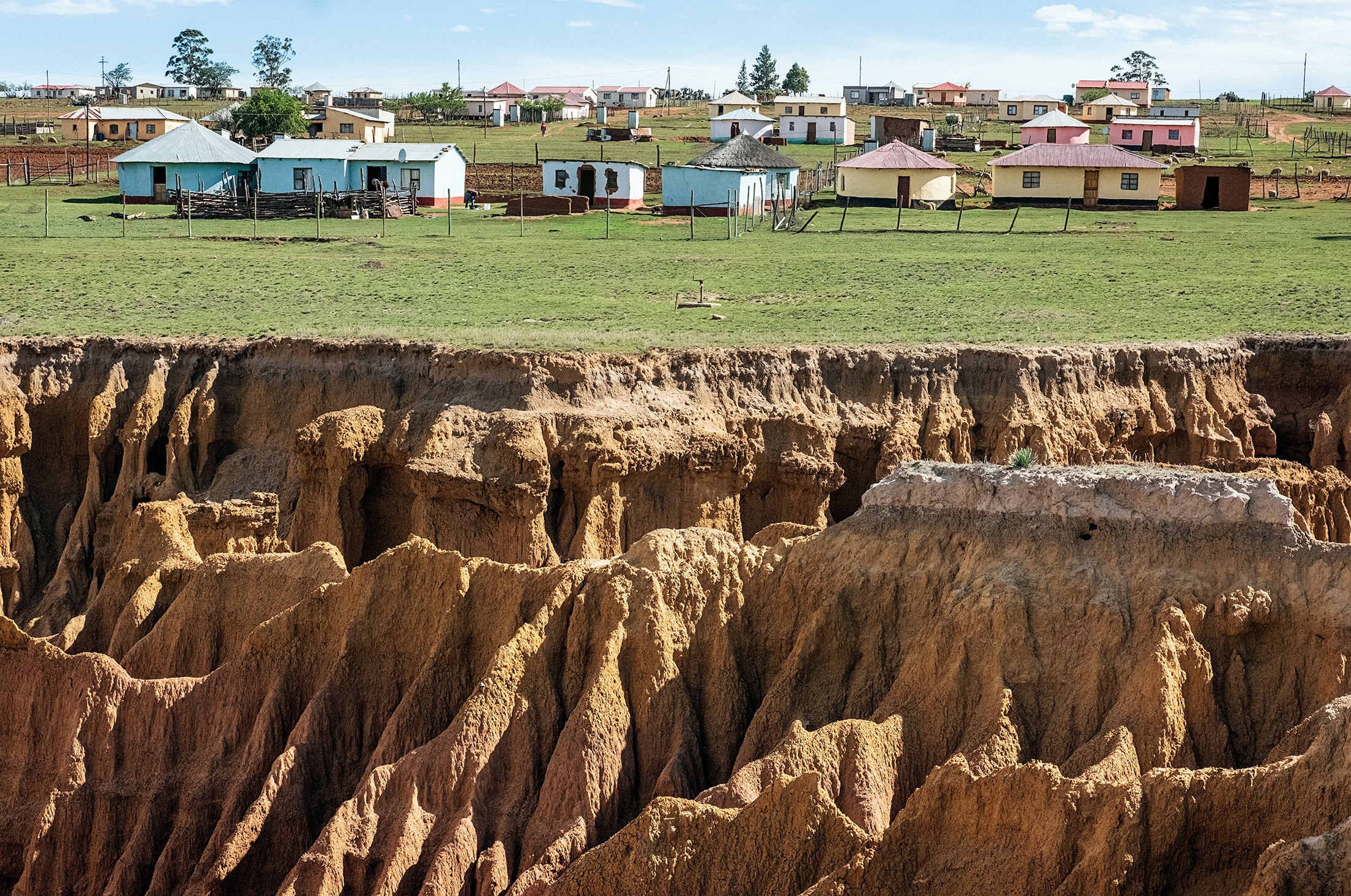 Lindokuhle Sobekwa's photograph 'Ezindongeni zase Kwezane' shows a settlement of houses in grassy field at the back, and eroded earth at the front.
