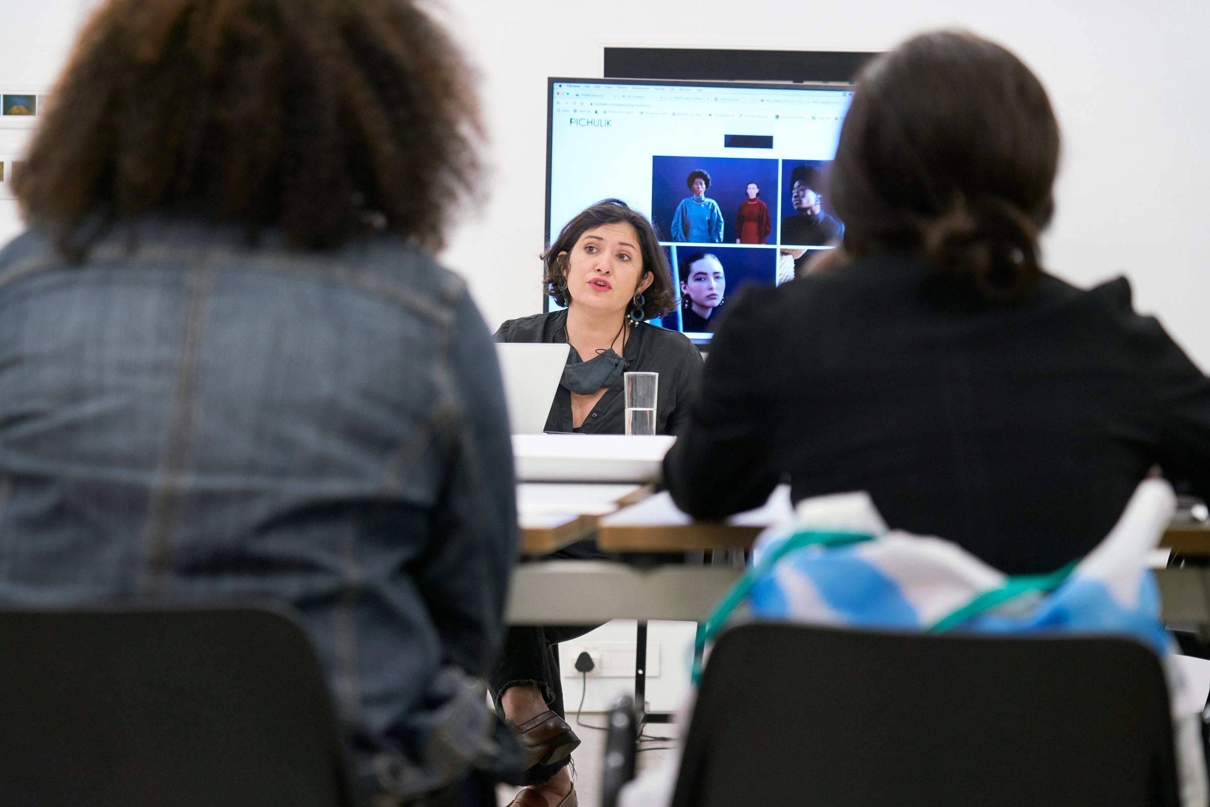 Event photograph from the Scalabrini Workshop that formed part of ‘Open Production’, Igshaan Adams’ hybrid studio/exhibition in A4’s Gallery. At the back, facilitator Kat Pichulik sits in front of a screen. In the front, attendees sit facing the facilitator.
