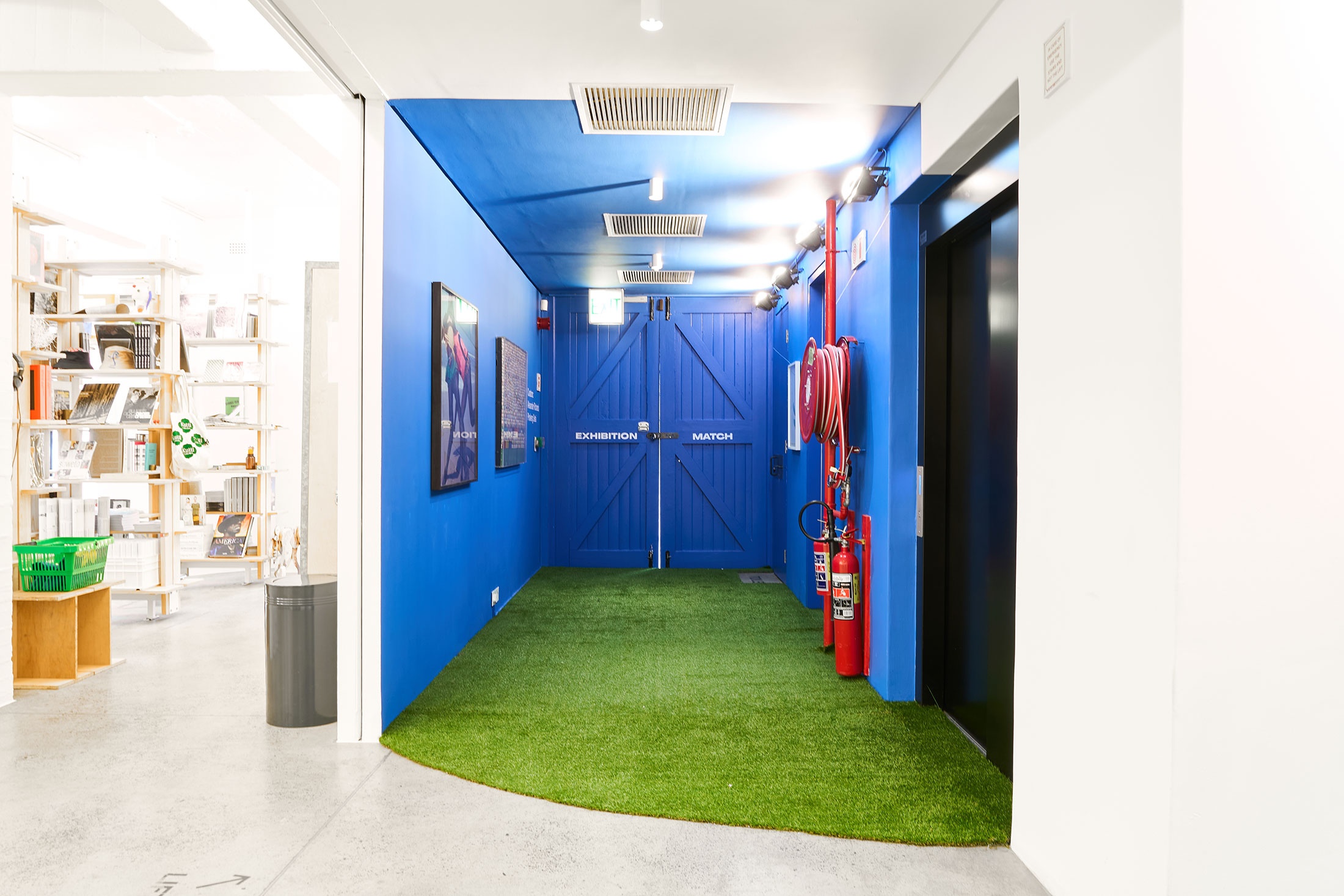 Installation photograph from the 2022 rendition of ‘Exhibition Match’ in A4’s Goods entrance. The floor is covered with artificial grass, with Viviane Sassan's photograph ‘La Lutte #2’ and Dan Halter’s mixed media work ‘Study with Colour (Dulux Range)’ on the lefthand wall.
