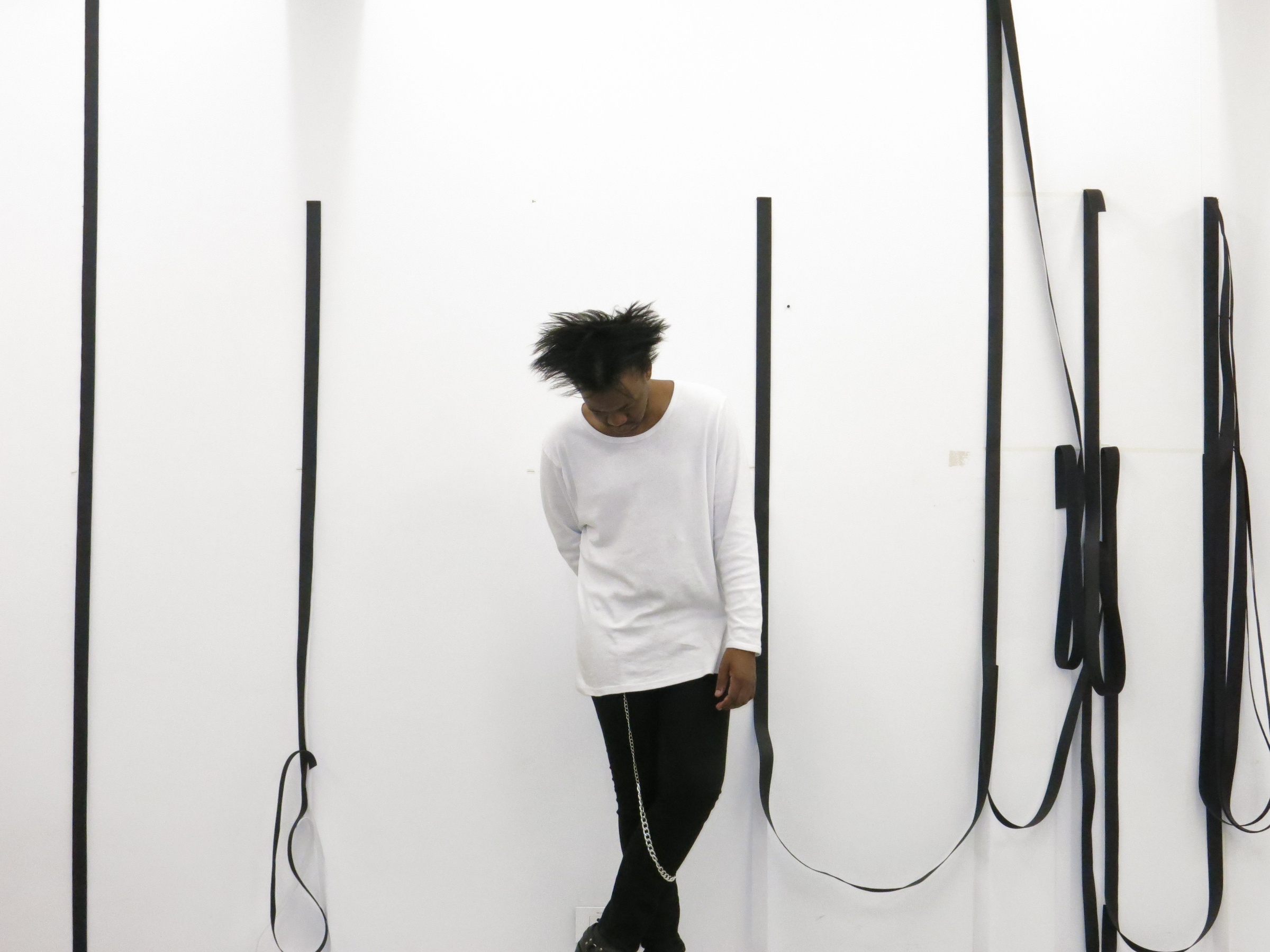 Process photograph from Unathi Mkonto’s interactive installation ‘AVOID’ on A4’s ground floor. At the back, strips of black velcro are arranged along the white wall. At the front, Mkonto stands looking downward.
