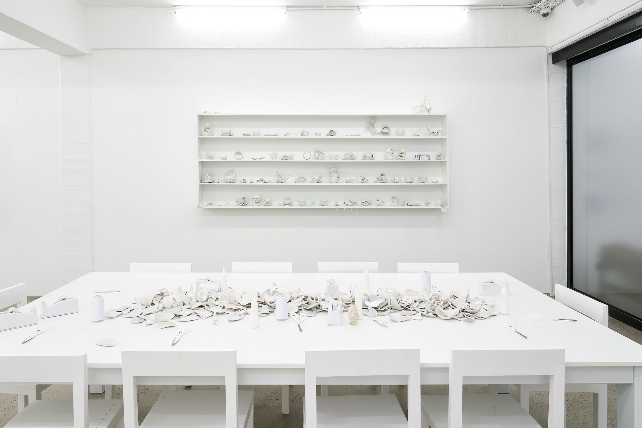 Installation photograph from the Customs exhibition in A4’s Gallery that shows Yoko Ono’s installation ‘MEND PIECE, A4 Arts Foundation, Cape Town version’ in A4’s ground-floor Reading Room. At the front, a white rectangular table with chairs holds ceramic fragments, tape, twine and scissors. At the back, a white wall-mounted shelving unit lines the white wall.
