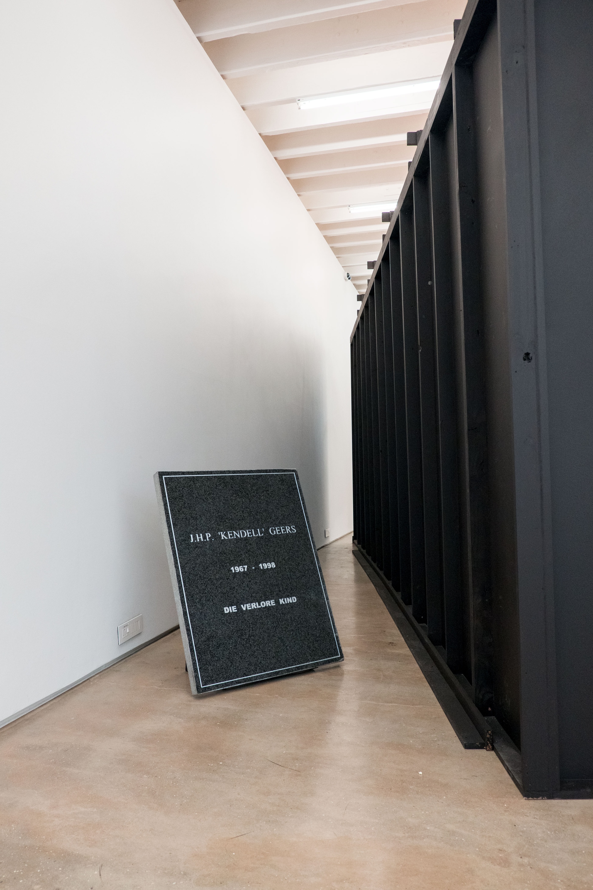 A photograph of Avant Car Guard's 'Bury J.H.P. “Kendell” Geers, 1967-1998 / Die Verlore Kind' shows a graphite slab with a white enamel inscription.
