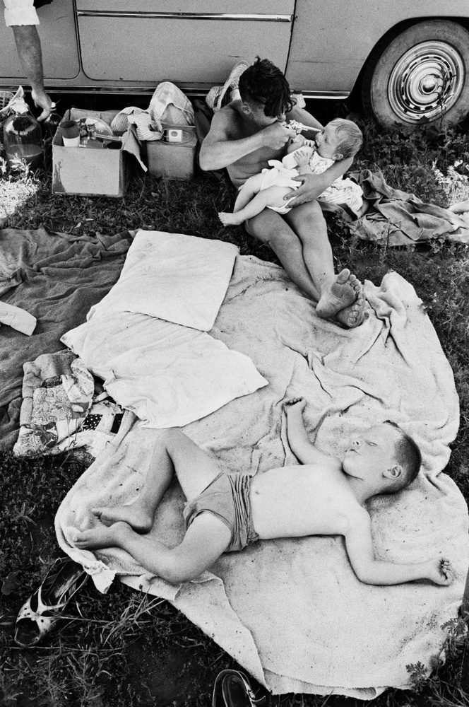 David Goldblatt's monochrome photograph 'Picnic at Hartebeespoort Dam on New Year’s Day, Transvaal (North-West Province)' shows two children on a picnic blanket in front of a car. The child at the back is holding a baby and pointing a toy gun at them.

