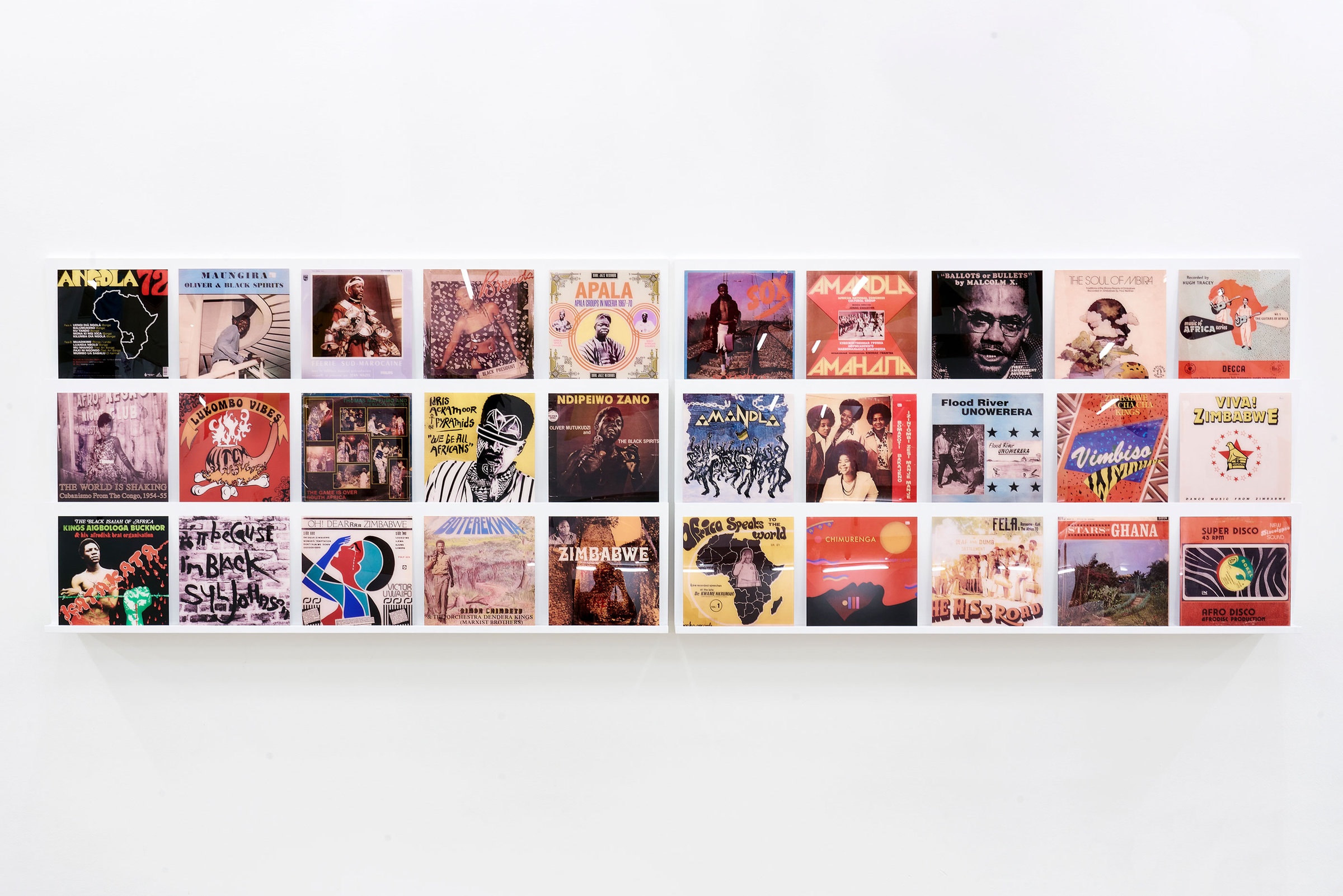 Installation photograph of Common exhibition. A grid of vinyl record cover reproductions from 'The Library of Things We Forgot to Remember’ by Kudzanai Chiurai is arranged on a slanted white shelf on a white wall.
