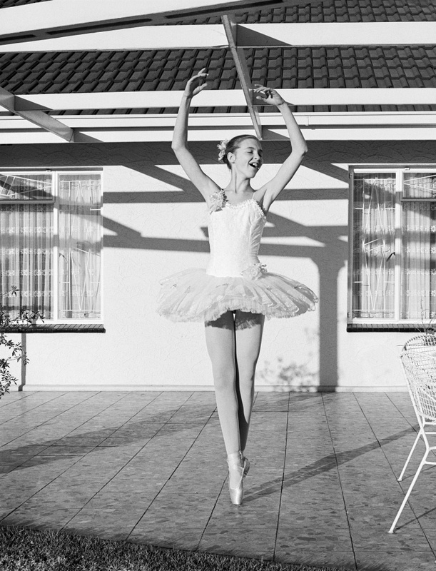 David Goldblatt's black-and-white photograph 'Girl in her new tutu on the stoep' (1980) shows a girl in a white leotard and tutu practising ballet outside. Behind her are two windows and shadows cast onto the wall by the rafters.
