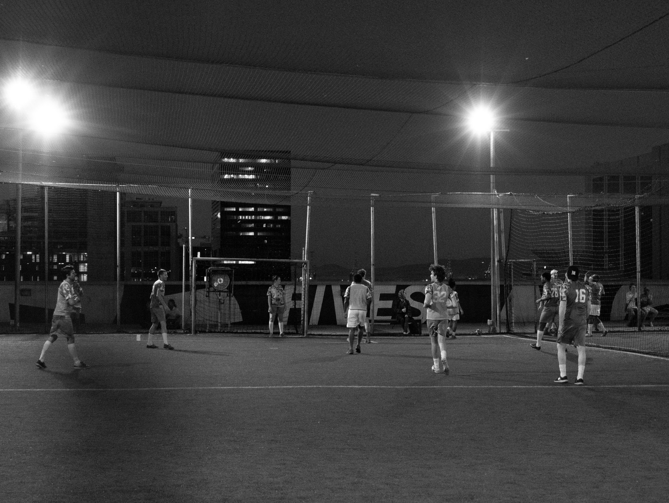 Monochrome event photograph from the 2023 edition of Exhibition Match depicts participants in a soccer match on a lit rooftop field at night.
