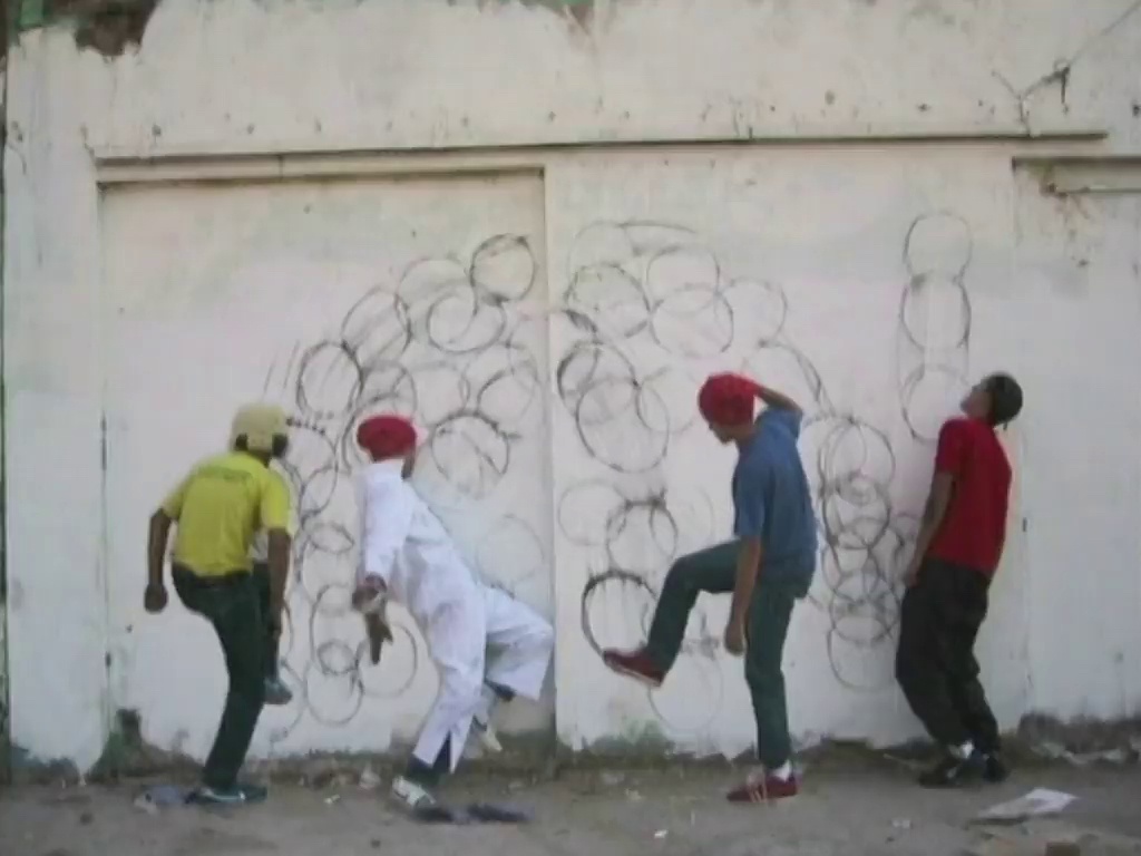 A still frame from Robin Rhode's digital animation 'Hondjie' shows four figures in motion with multiple drawings of soccer balls on the wall behind them.
