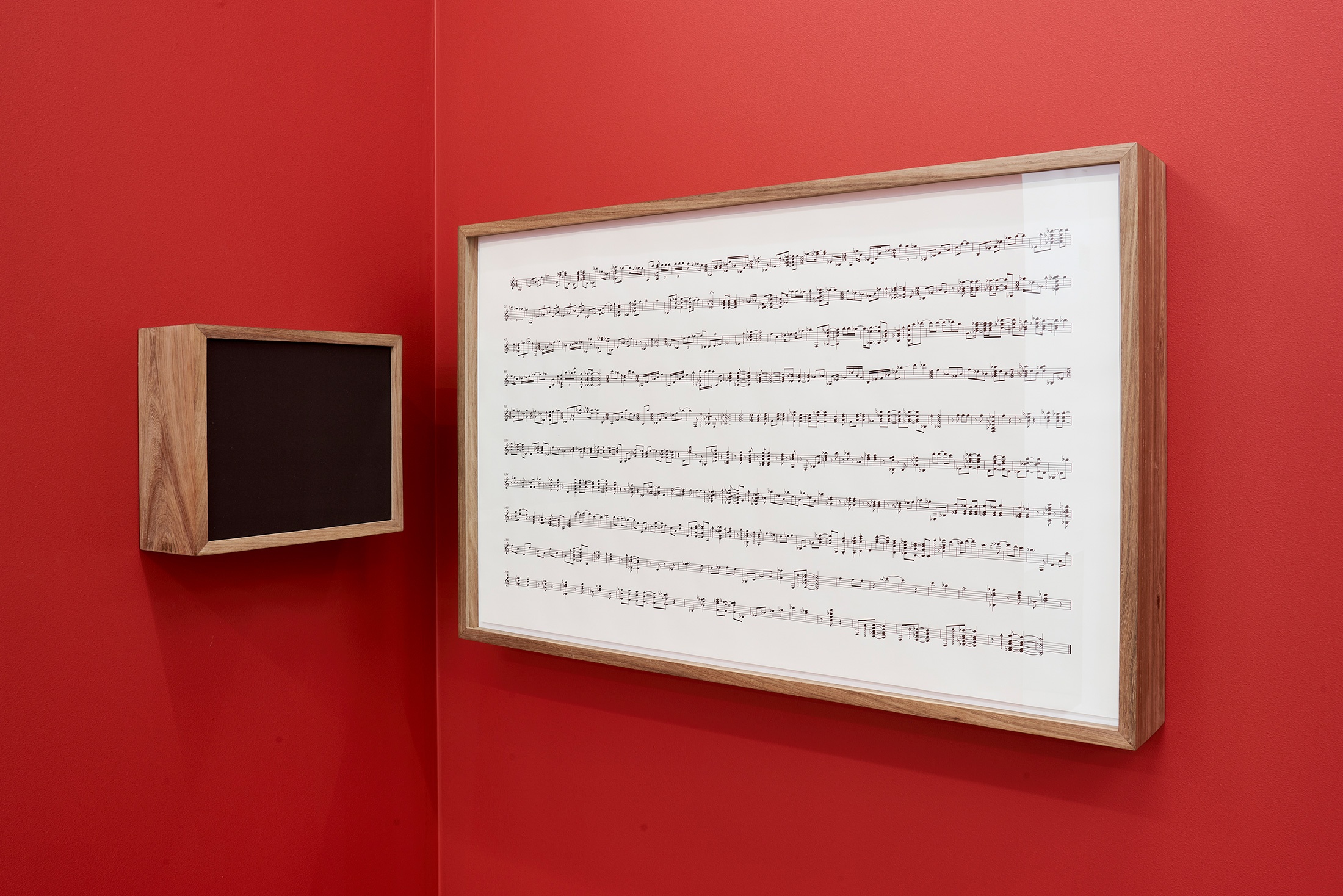 Installation photograph that shows Iñaki Bonillas’ wall-mounted installation ‘The Return to the Origin 7’ in the corner of two red movable gallery walls. On the left is a wood-framed speaker box and on the right, a wood-framed musical score.
