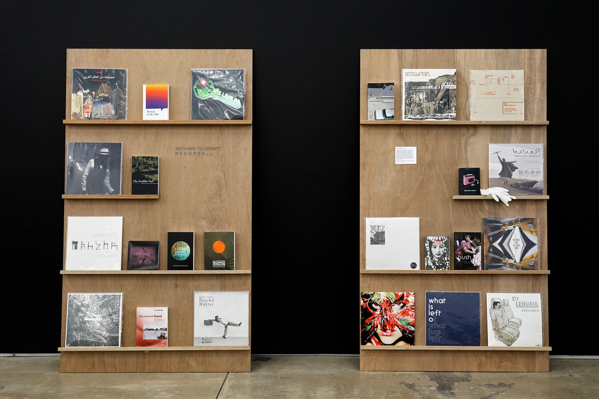 Installation photograph from the ‘Sounding the Void, Imaging the Orchestra V.1’ exhibition in A4’s Gallery. A selection of vinyl records from Bhavisha Panchia’s ‘Nothing to Commit Records’ collection and some printed matter is arranged on two rectangular wooden stands with narrow shelves.
