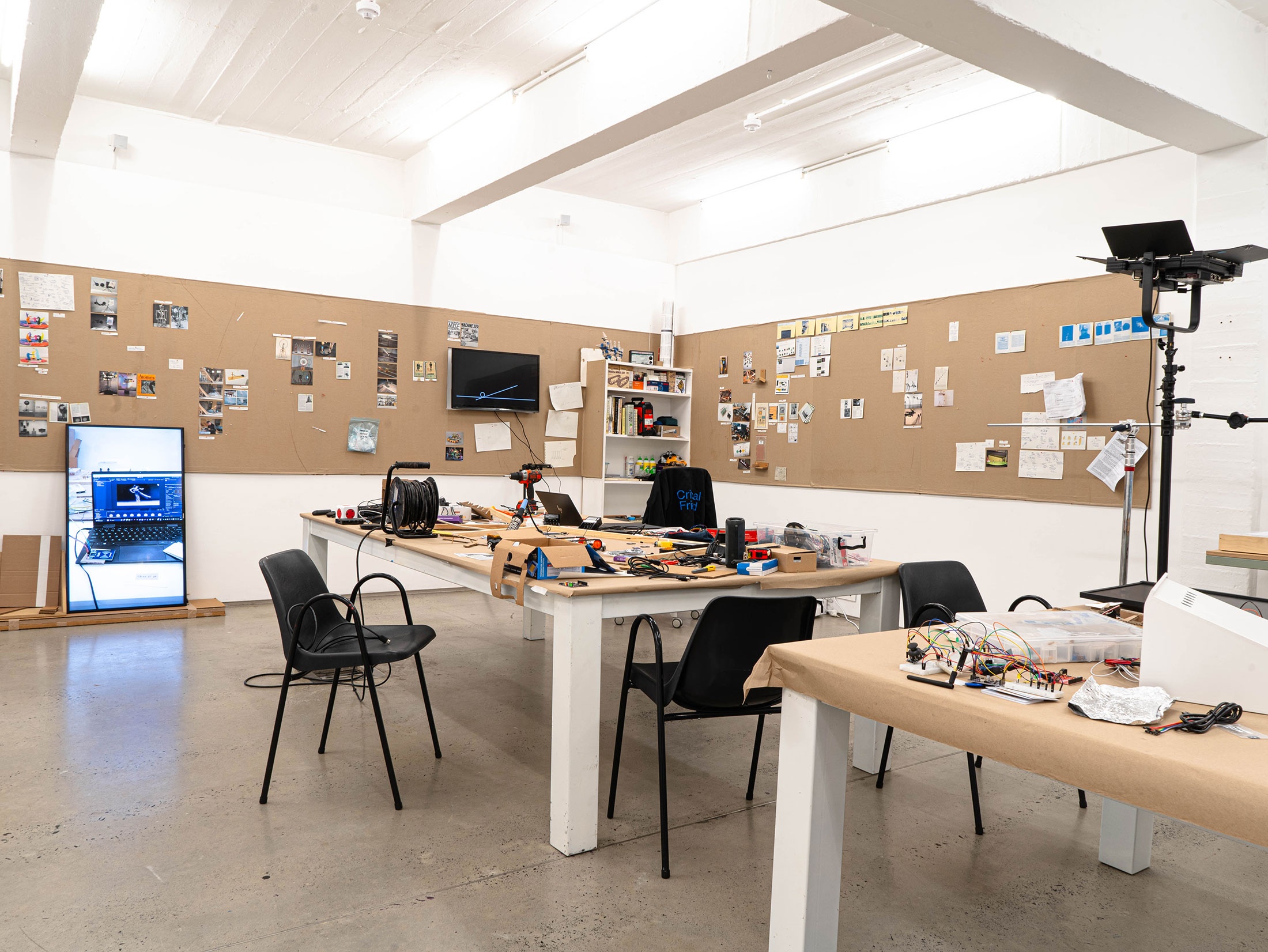 Installation photograph from Mitchell Gilbert Messina's residency in A4 Art Foundation. In the middle, two tables wrapped in brown paper holds pieces of cardboard and various electronic tools and components. At the back, the walls lined with pieces of cardboard that host pinned research notes.
