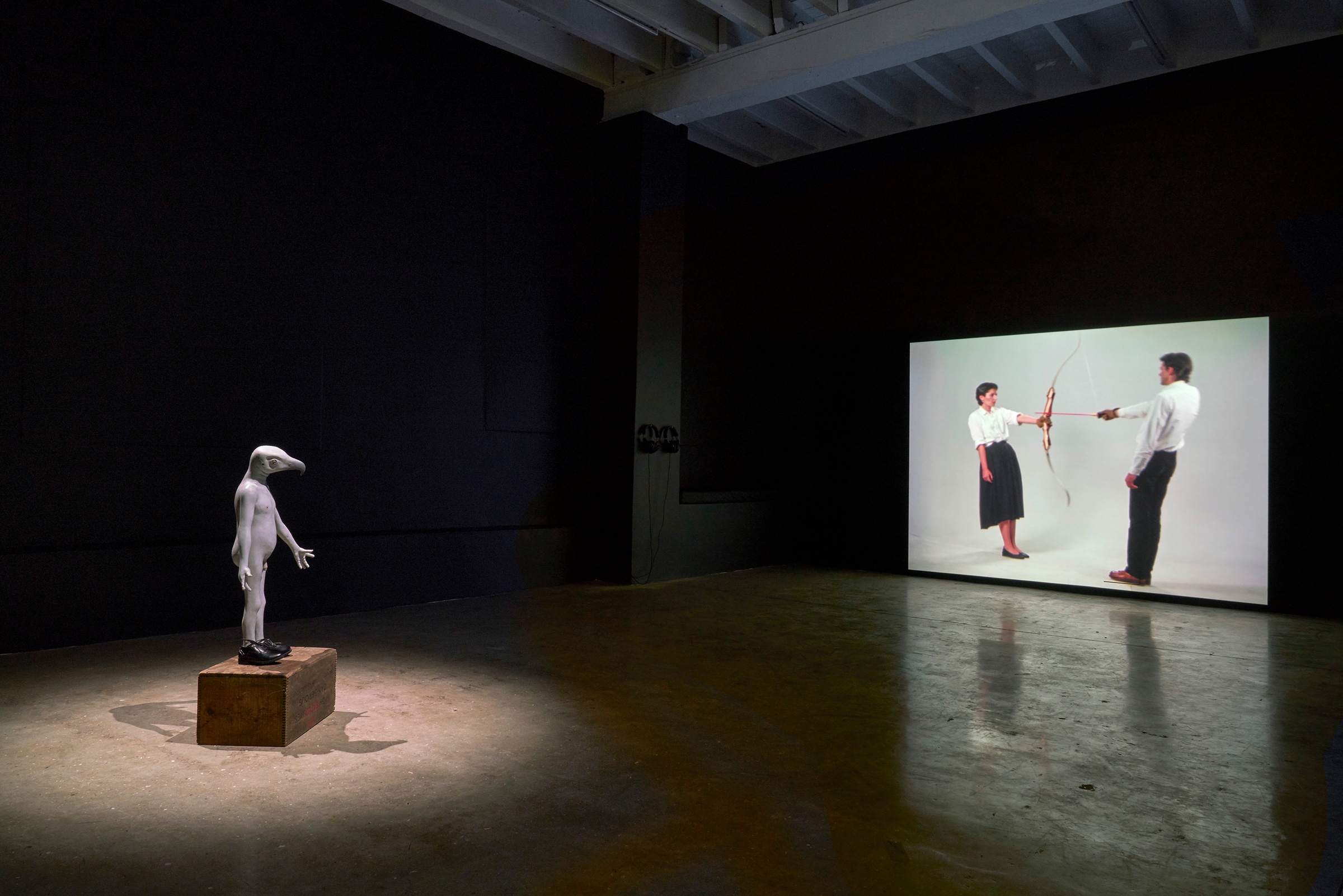 Installation photograph from the ‘Risk’ exhibition in A4’s Gallery. On the left, Jane Alexander’s sculpture ‘Security Bird’ sits under a spotlight. On the right, Ulay and Marina Abramović’s performance video ‘Rest Energy’ is projected onto the wall.
