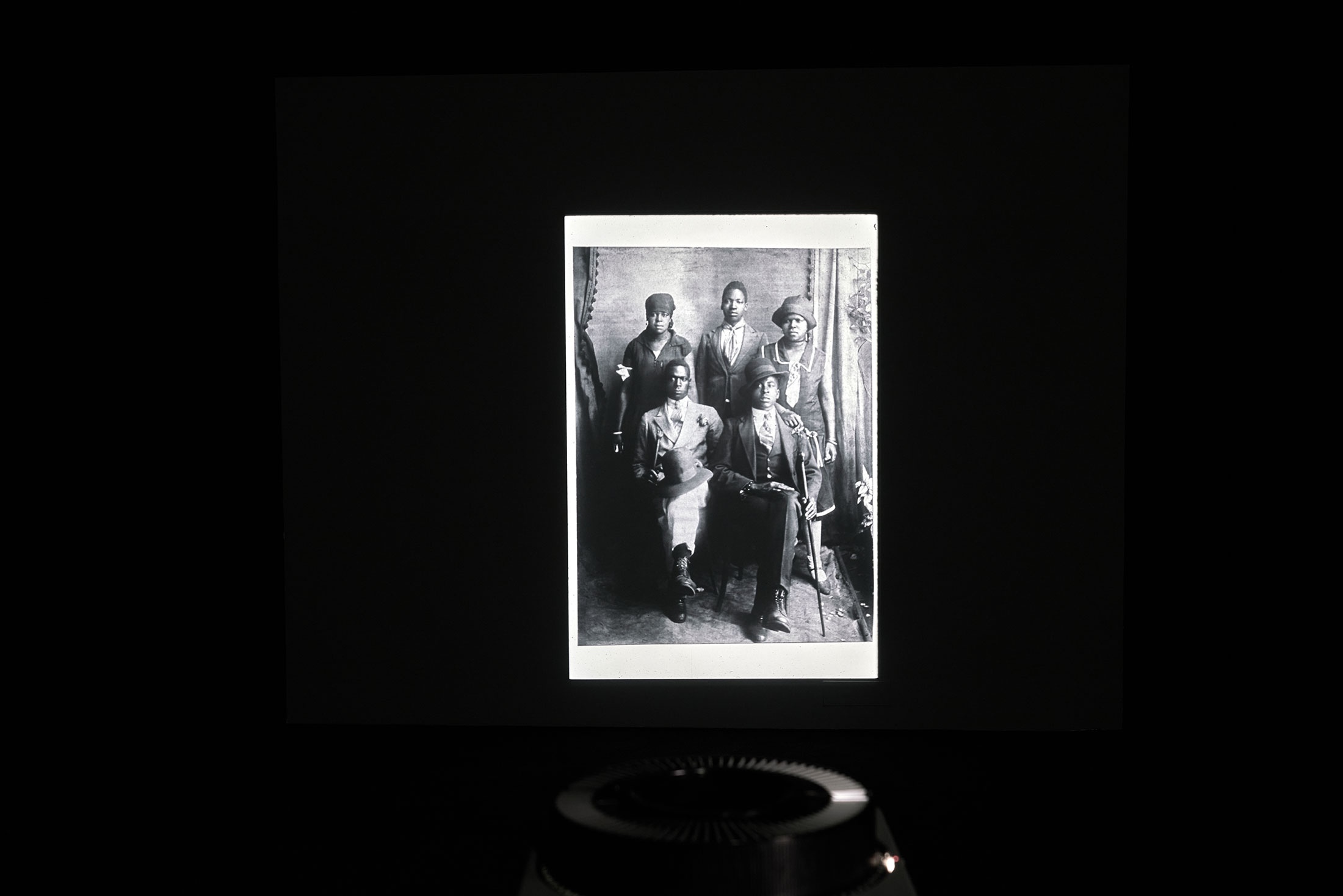 A photographic slide from Santu Mofokeng's 'Black Photo Album/Loot at Me' shows two individuals seated on a wooden bench looking at the photographer.
