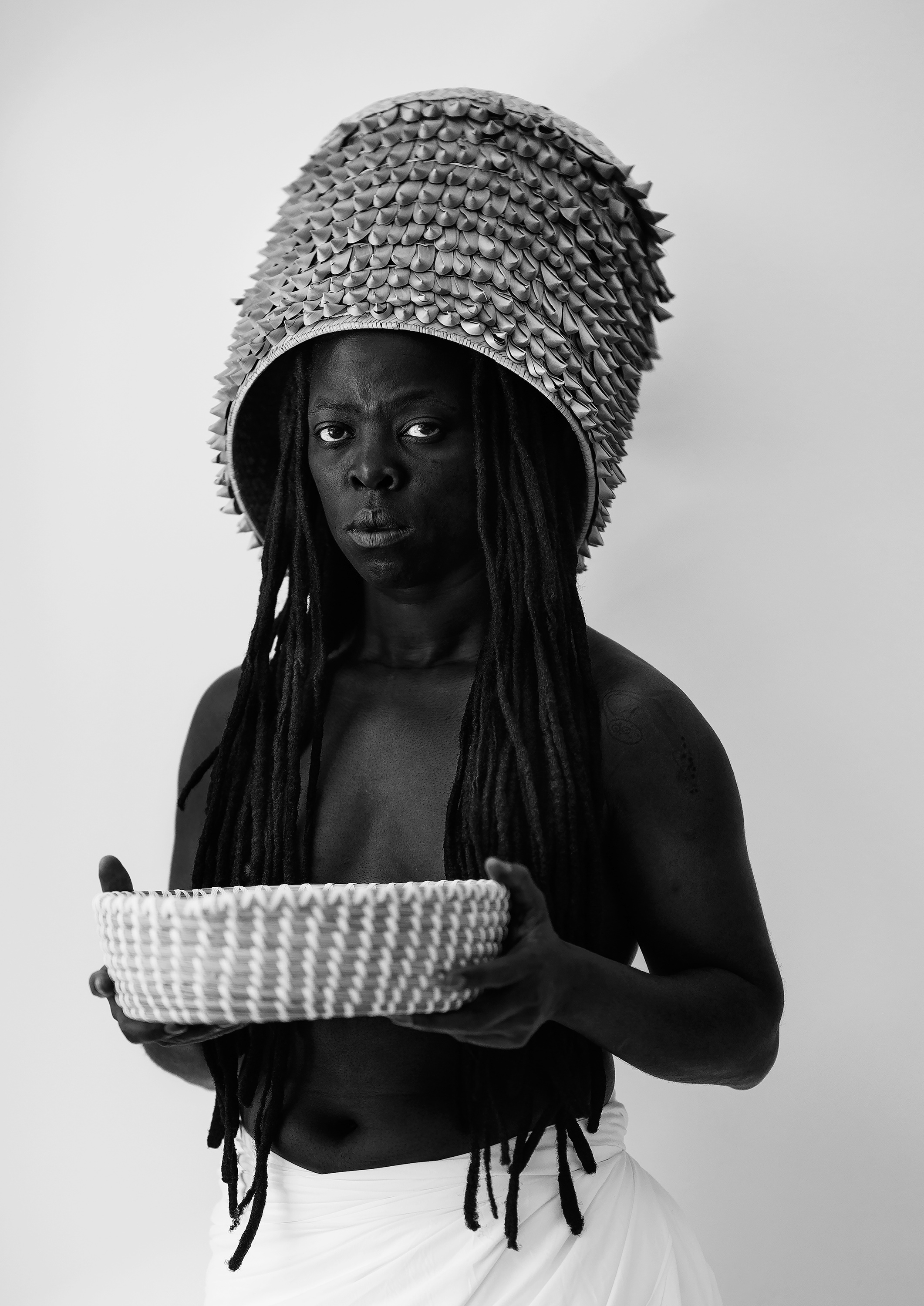 Zanele Muholi’s monochrome photograph ‘Hlanzeka III, Vineyard Hotel, Room 153, Cape Town, 2017’ shows the artist standing in front of a white backdrop holding a basket.
