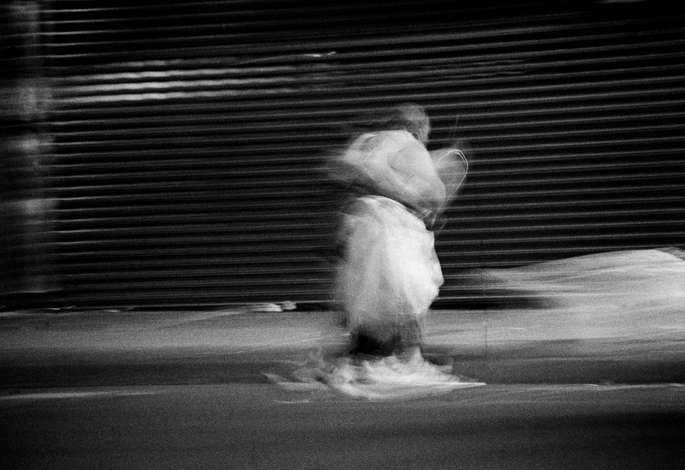 Sabelo Mlangeni's monochrome photographic print 'Invisible Woman III' shows a woman walking on a concrete sidewalk.
