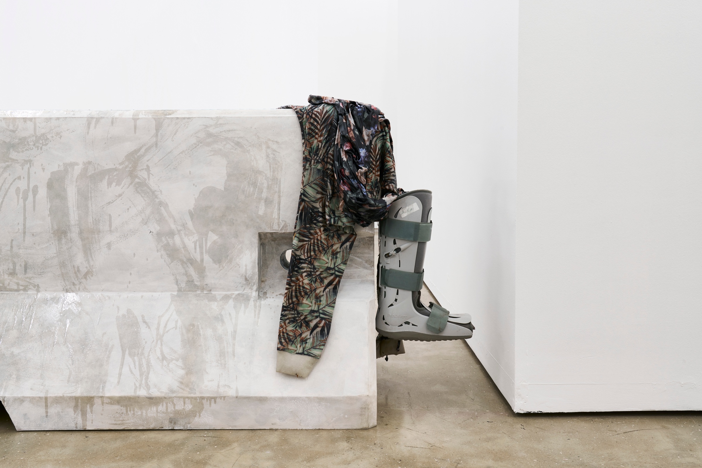 A photograph of Kevin Beasley's untitled plywood, paint, resin, housedress, sweatshirt and walking boot sculpture sitting on a concrete floor.
