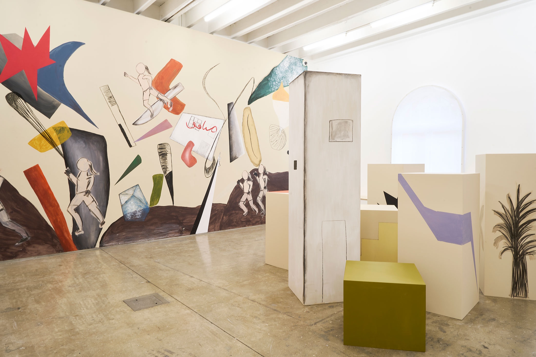 Installation photograph of the Common exhibition, featuring an installation by Hanna Noor Mahomed called ‘The Prodigal Daughter.’ On the right, a group of plinths painted with geometrical shapes and objects to resemble a city. At the back, a wall mural with four femme figures running amongst various objects and geometrical shapes.
